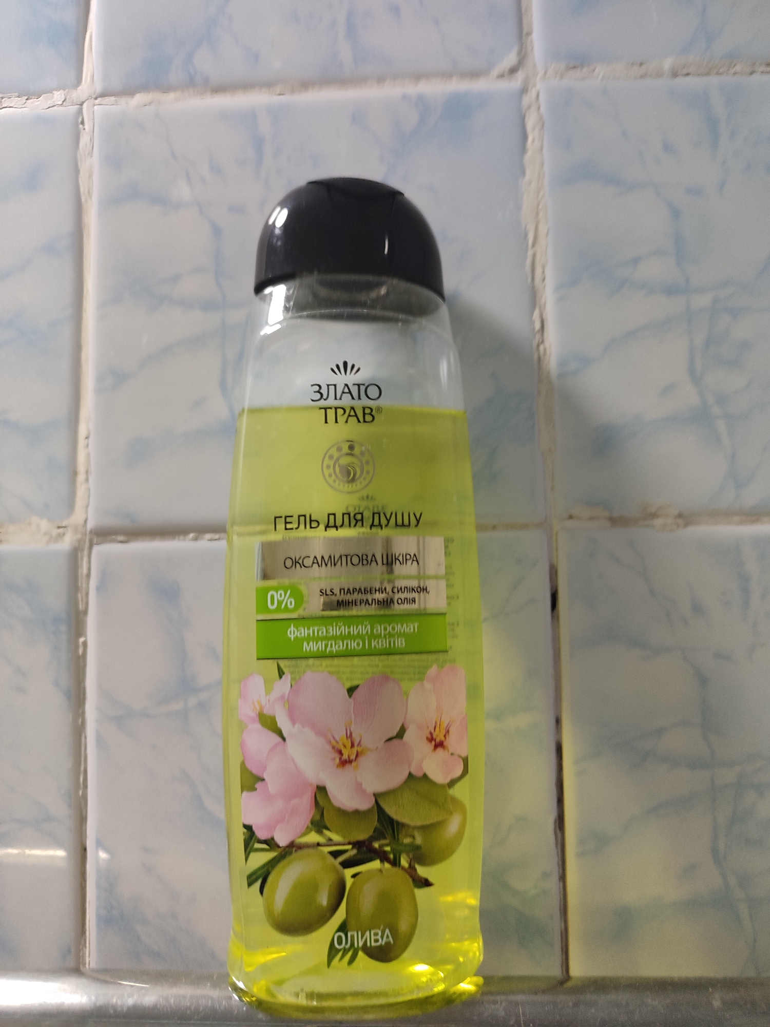 A clear plastic bottle of shower gel with a black cap stands on a metallic surface. The liquid inside is a light yellow color. The bottle has a label with text in Ukrainian, featuring a logo, the words "ЗЛАТО ТРАВ" and "ГЕЛЬ ДЛЯ ДУШУ," followed by "ОКСАМИТОВА ШКІРА." Below this, it states "0%" and lists "SLS, ПАРАБЕНИ, СИЛІКОН, МІНЕРАЛЬНА ОЛІЯ." Further down the label is "фантазійний аромат мигдалю і квітів" and an image of pink flowers and green olives. The background consists of light blue and white tiled walls, which appear to be in a bathroom setting. The scene is likely located within the city of Mykolaiv, Ukraine, given the Ukrainian text on the product.