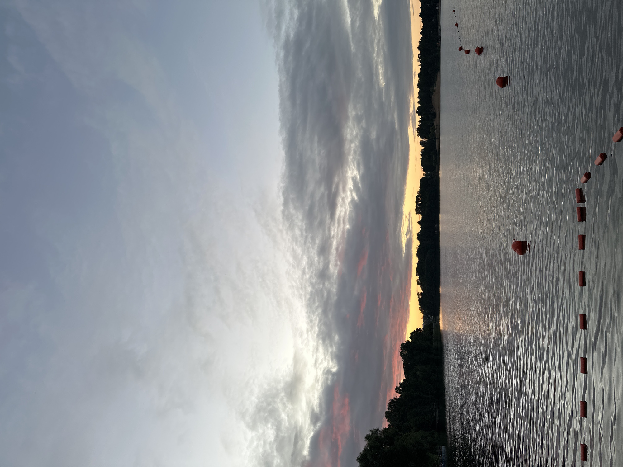 The image depicts a serene lake scene at dusk with a softly colored sky. In the foreground, a line of orange buoys stretches across the rippling water, marking a section of the lake. Further out, a single larger orange buoy floats. The far bank of the lake is lined with a dense silhouette of trees. The sky above is filled with dramatic clouds, transitioning from a pale blue at the top to hues of pink and orange near the horizon, suggesting the last moments of sunset. The overall atmosphere is tranquil and natural. The image does not provide any information about the city of Saint Petersburg, Russia, or any specific location.