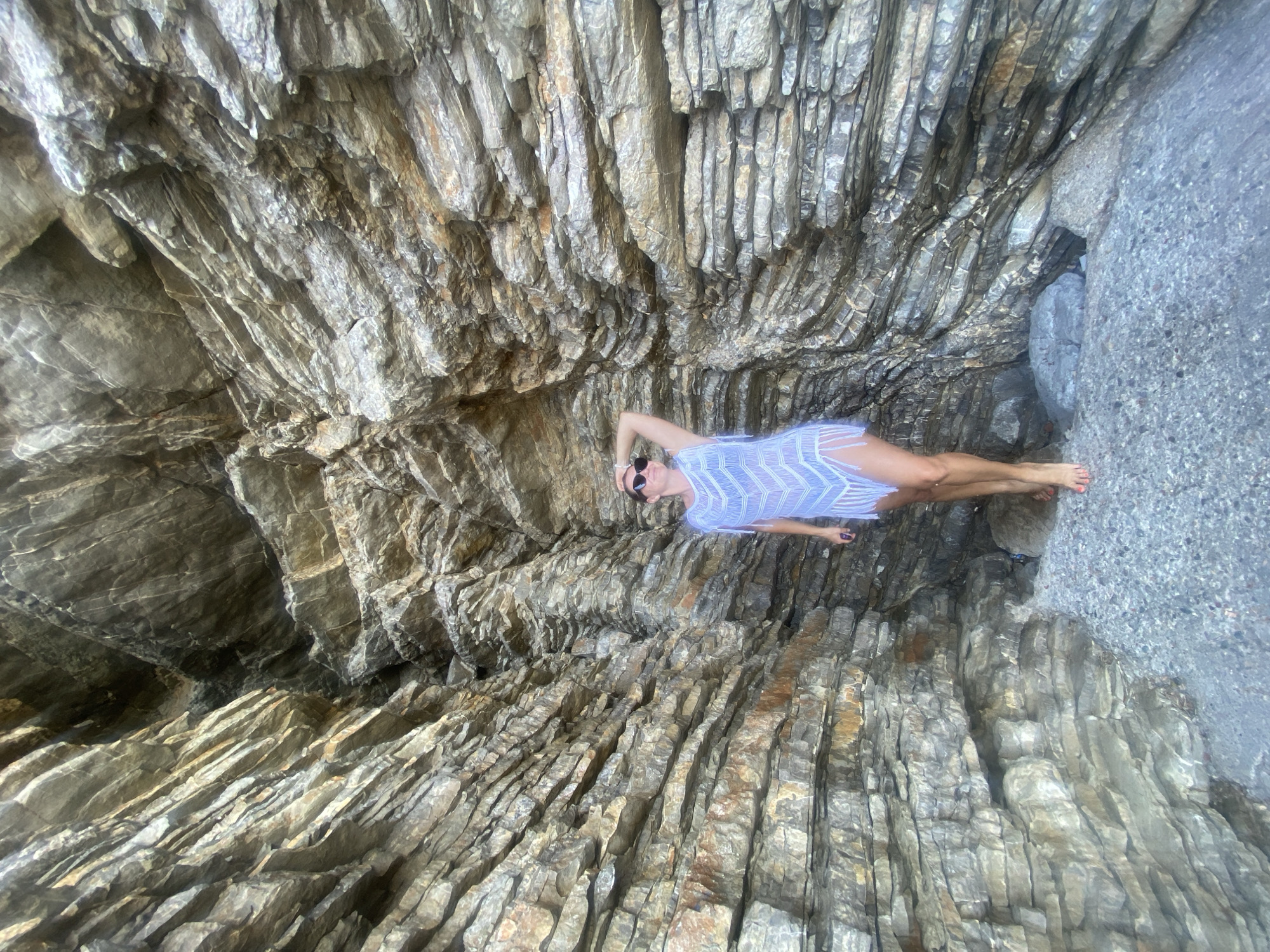 A woman stands between two rock formations on a beach. She is wearing a white, fringed dress and sunglasses. The rocks are layered and have a rough texture, with a mix of grey, brown, and tan colors. The ground is covered in small stones and pebbles. The scene appears to be in a natural cove or grotto, likely on the coast of Pržno, Montenegro, given the characteristic layered rock formations often found in the region. The lighting suggests it is daytime, with natural sunlight illuminating the scene.