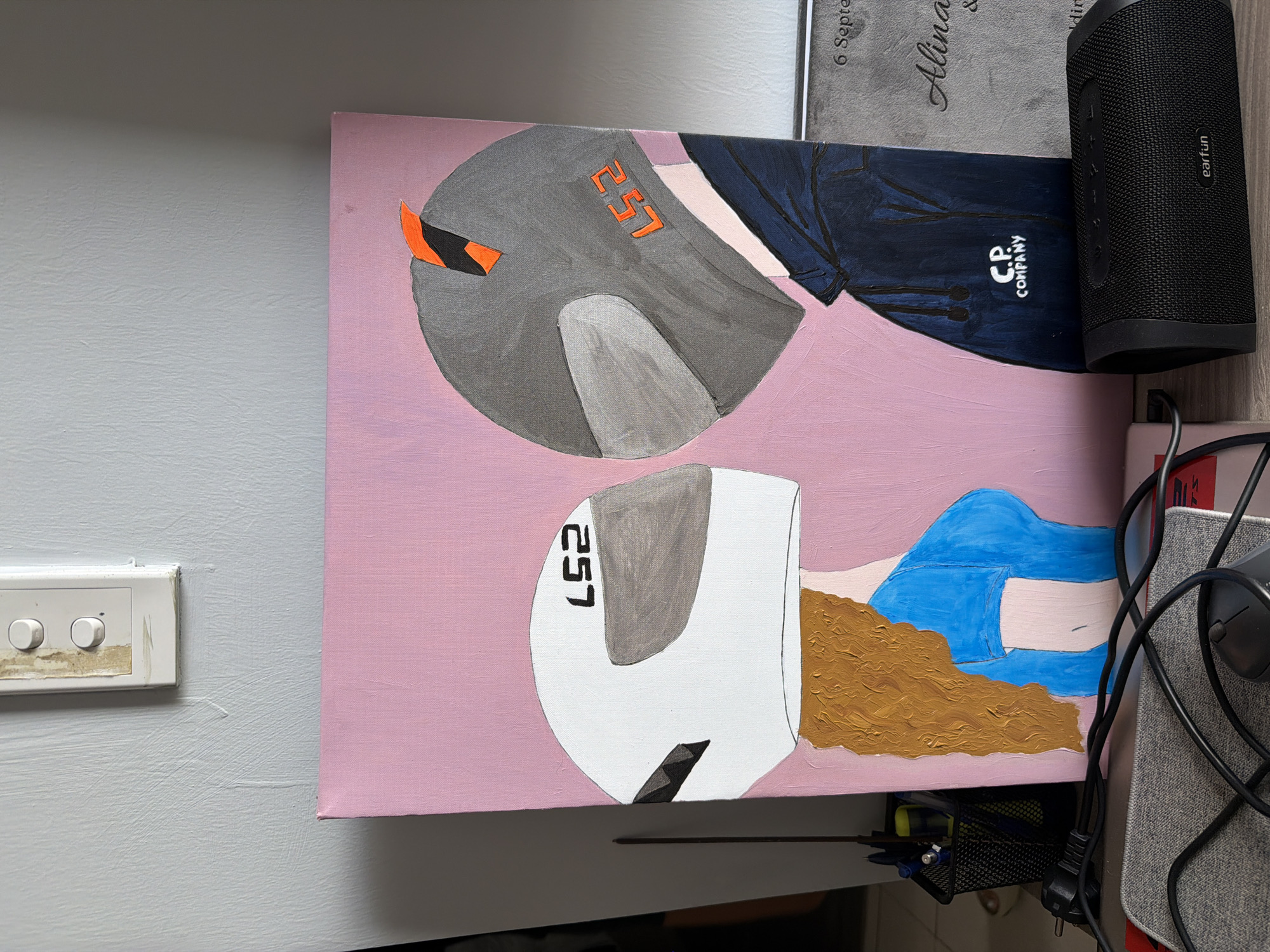 The image shows a painting of two people wearing motorcycle helmets. The painting is on a pink canvas and is leaning against a wall. To the left of the painting is a black wire basket filled with pens and markers. In front of the painting is a black portable speaker with the brand name "earfun" visible. A grey mouse and black cables are also present in the foreground. The wall behind the painting has a light switch with two buttons. The scene appears to be indoors. There is no information in the image to suggest the location is in Nha Trang, Vietnam.