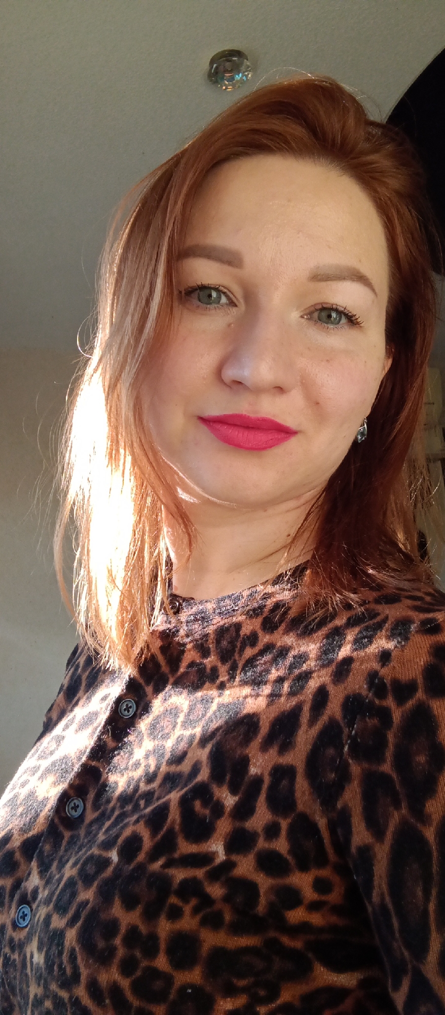 A woman is centered in the image, cropped at the mid-chest level. She has fair skin, light green eyes, and red hair that is partially illuminated by sunlight on the left side of the frame. Her lips are painted with bright pink lipstick. She is wearing a dark brown and black leopard-print cardigan with visible buttons. A crystal light fixture is visible in the upper left of the frame. An earring is visible on the woman's left ear. The background is out of focus and indistinct. The location is Unknown, Unknown.