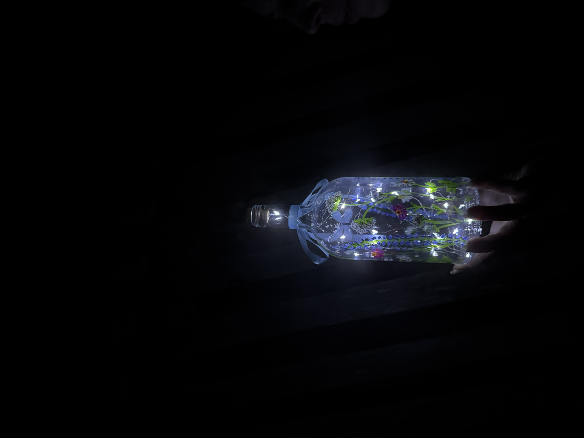 A horizontally-oriented, close-up shot depicts a glass bottle, possibly containing fairy lights and floral elements, being held in the dark by a hand. The bottle is the main source of light in the image, illuminating the hand holding it. The background is completely dark, except for the vague shape of what seems to be a person's face at the top of the frame, slightly above the bottle. The location is not discernible from the image.
