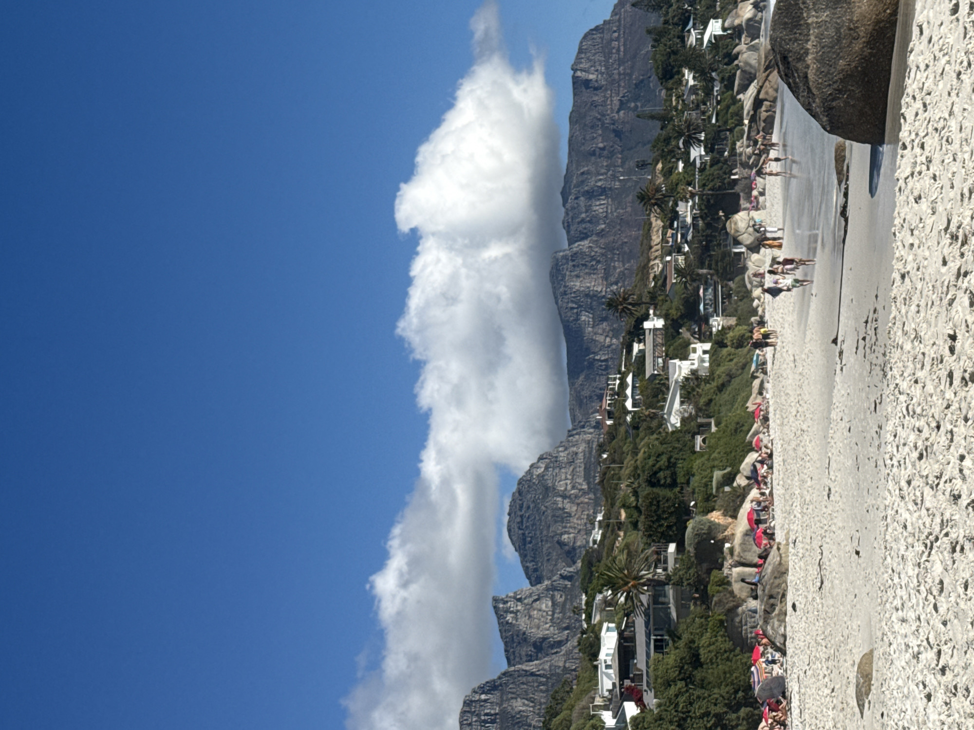 The image captures a scene in Cape Town, South Africa, featuring a beach with white sand and large boulders. People are present on the beach, some walking along the shoreline and others seemingly relaxing. Buildings and greenery are visible on the hillside behind the beach. A large cloud formation extends vertically, partially obscuring the mountain range in the background. The sky is bright blue.