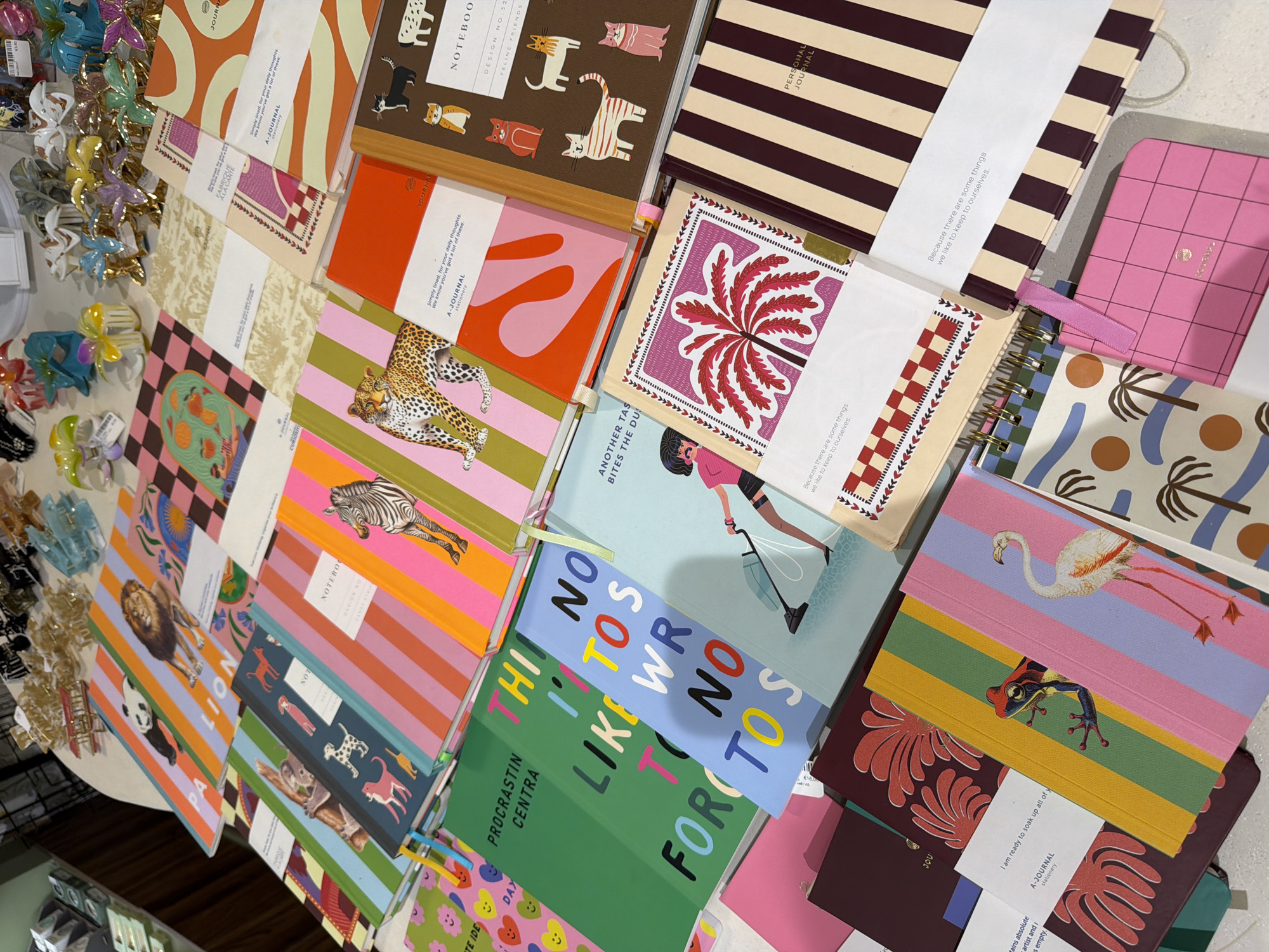 This image displays a collection of vibrant and artistically designed notebooks and journals, interspersed with colorful hair claws. The subjects are primarily inanimate objects, with notebooks featuring playful illustrations of animals like cats, lions, pandas, zebras, and koalas, as well as abstract patterns and botanical designs. Some notebooks have text printed on their covers, including titles like "NOTEBOOK," "PERSONAL JOURNAL," "FELINE FRIENDS," and motivational phrases such as "Simply lined, for your daily thoughts" and "We know you've got a lot of these." Other notebooks have humorous or whimsical slogans like "PROCRASTIN CENTRA" and "ANOTHER TAS BITES THE DU". The hair claws, in various bright colors like pink, blue, yellow, and purple, are scattered among the notebooks, adding a playful and somewhat chaotic element.
The scene appears to be an indoor retail setting, possibly a stationery shop or a display area, with a white countertop or table surface visible. The lighting suggests it could be daytime, though no specific time of day can be definitively determined. There are no people or animals actively engaged in the scene, and no weather cues are present. The overall impression is one of a cheerful and creative display of stationery items. The location context of Mdina, Malta, does not offer any specific visual cues within the image itself to confirm or deny this setting.