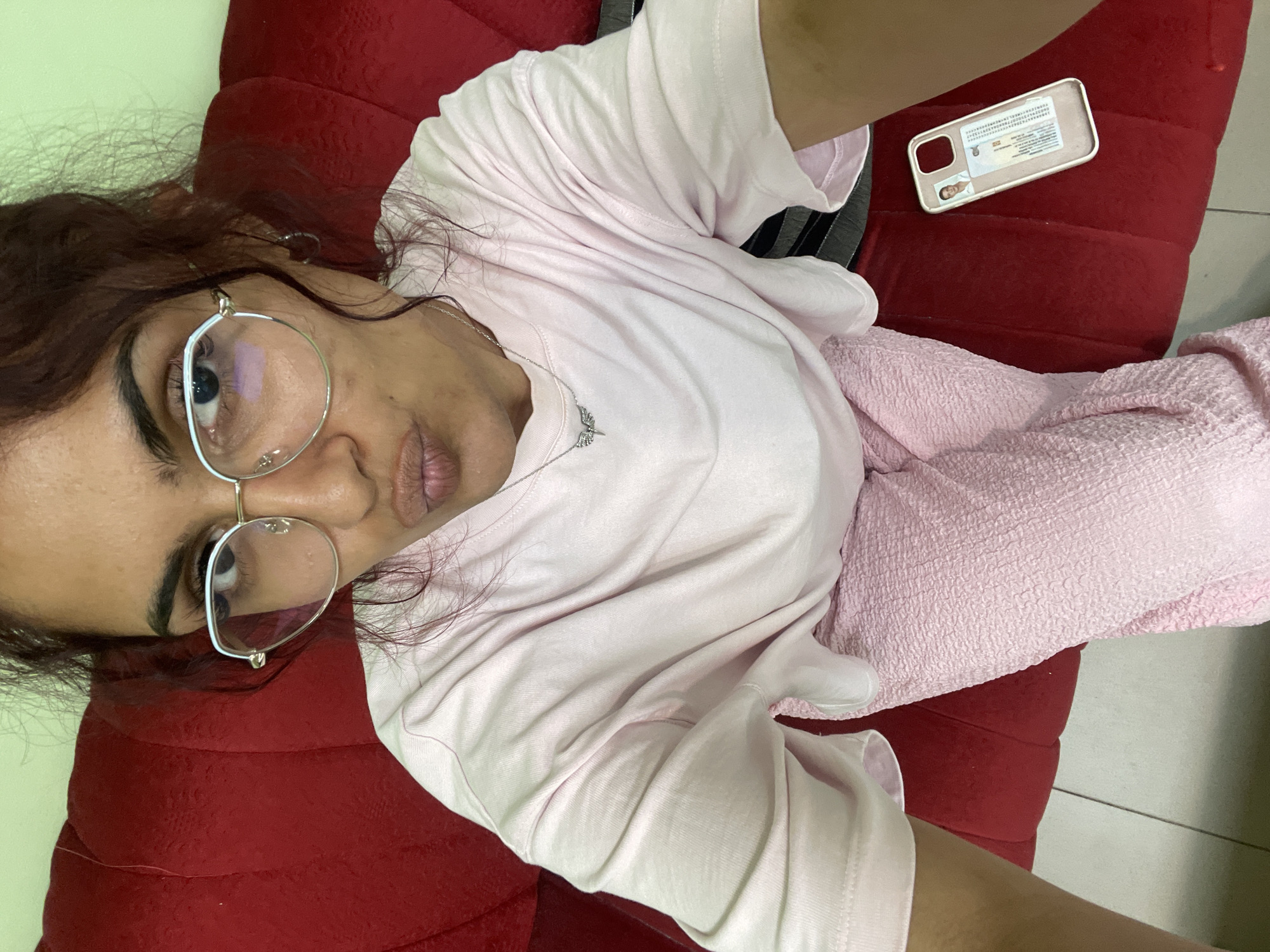 A high-angle selfie captures a person with medium-to-dark skin tone, dark eyebrows, and wavy, dark reddish-brown hair, wearing round, light-framed eyeglasses. The person has their lips puckered and is looking directly into the camera. They are wearing a light pink, short-sleeved t-shirt and light pink, textured pants. A silver-colored necklace with a wing-shaped pendant is visible, along with a silver hoop earring in the right ear. The person is seated on a plush, red-upholstered couch. To the right of the person's leg, on the red couch, lies a light-colored smartphone case containing a card resembling an identification document with a photograph of a male individual. The background features a light green wall behind the person's head and shoulders, and a light-colored tiled floor is partially visible at the bottom left. The location is an indoor setting in Unknown, Unknown.