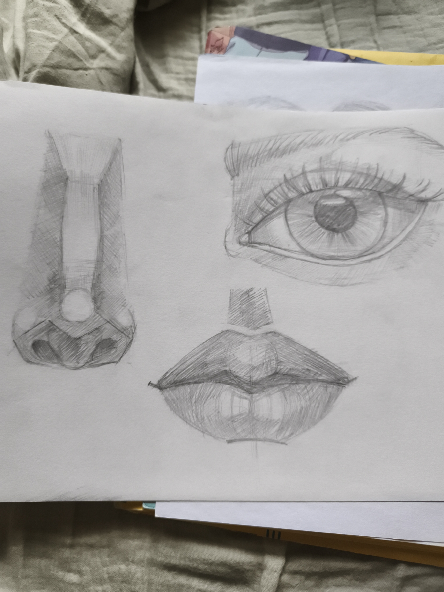 The image is a close-up, overhead view of a pencil sketch on a sheet of white paper. The sketch depicts parts of a human face: a nose on the left, an eye in the upper right, and lips in the lower center. The nose is drawn from the bridge down to the nostrils, with shading used to indicate form and shadow. The eye is also rendered with detail, showing the iris, pupil, and eyelashes, with careful attention to light and shadow. The lips are full and sensually drawn, with shading suggesting their shape and texture. The sketch is done on a flat surface, possibly a table or desk, with a wrinkled, textured fabric visible in the background. The lighting appears to be natural, casting soft shadows on the paper. There are no people, actions, or interactions depicted in the image. The location in the city of Saint-Denis, France, is not discernible from the provided image.