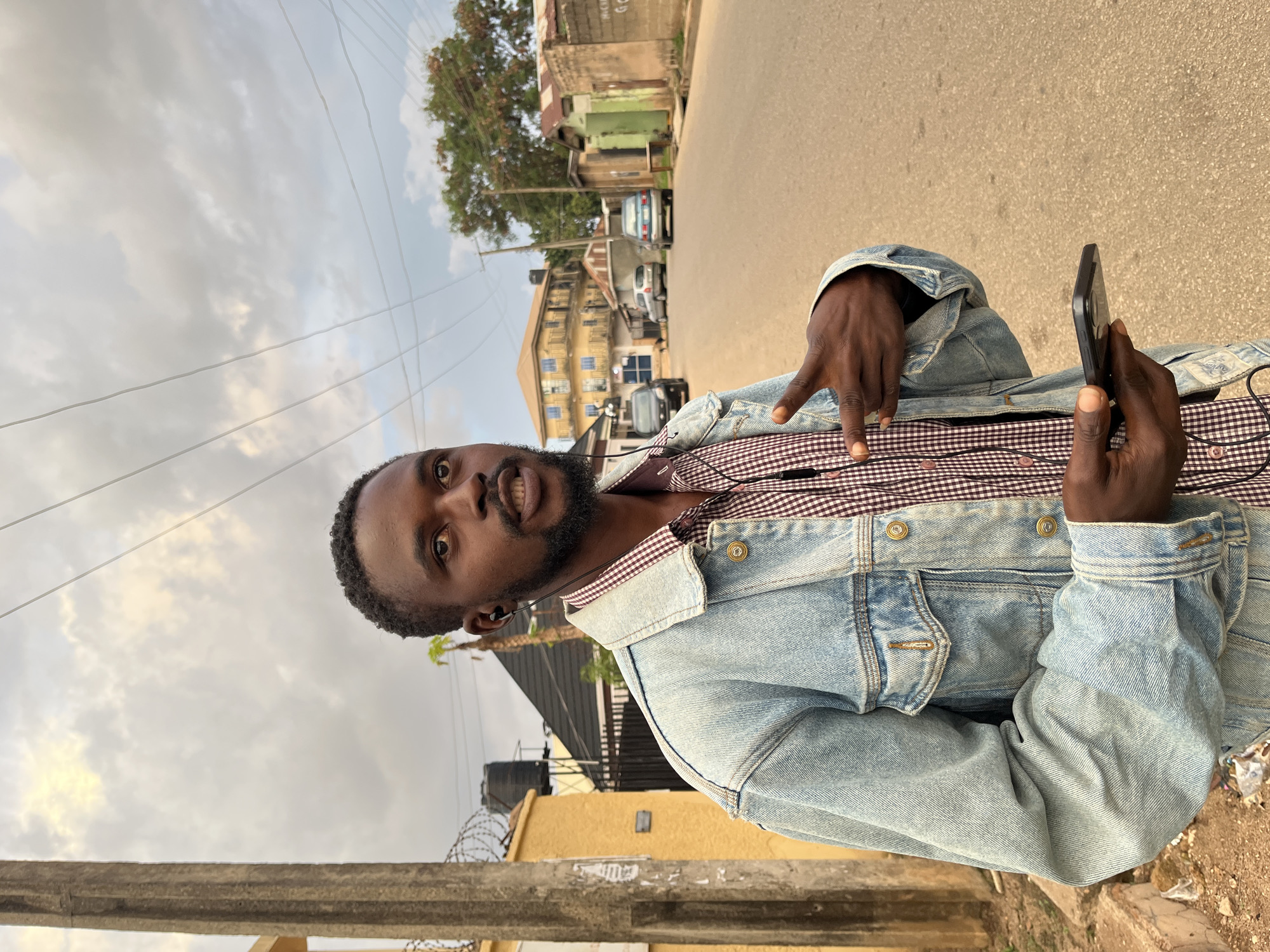 A young man with dark skin, a short beard, and closely cropped dark hair is captured in a full-length outdoor portrait, positioned on a street in what appears to be Jos, Nigeria. He is wearing a light blue denim jacket over a reddish-brown and white checkered shirt, and has earbuds in his ears. He looks directly at the camera with a slight smirk or open-mouthed smile, holding a black smartphone in his right hand. His left hand is raised, forming a 'V' shape with his index and middle fingers, possibly a peace sign or indicating the number two.
The background reveals a bustling street scene under an overcast or cloudy sky. Numerous power lines crisscross the sky. Along the street, various buildings of different sizes and states of repair are visible, some appearing weathered, with parked cars scattered along the roadside. A tall tree with green foliage is prominent in the middle ground, near some of the buildings. A concrete barrier or low wall occupies the foreground. The ground appears to be a mix of paved and dusty areas. The overall atmosphere suggests a casual daytime moment in an urban or semi-urban setting. No discernible text is visible in the image.