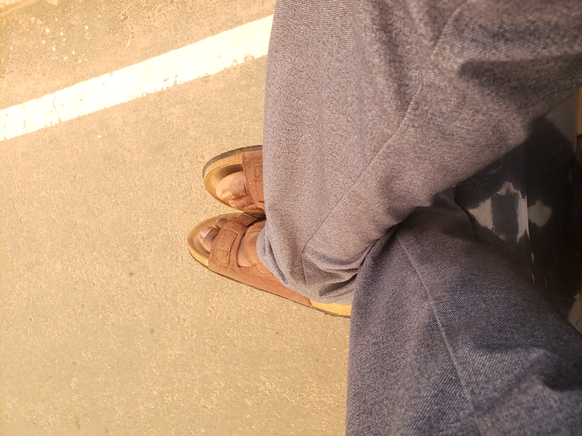 The image shows a person's legs and feet, clad in brown sandals and dark grey pants, sitting on a dark surface. The ground is a light brown, gritty material with a faint white stripe running diagonally across it, suggesting an outdoor setting, possibly a road or sidewalk in Narayi, Nigeria. The sunlight casts a bright reflection on the ground, and the overall lighting suggests it is daytime.