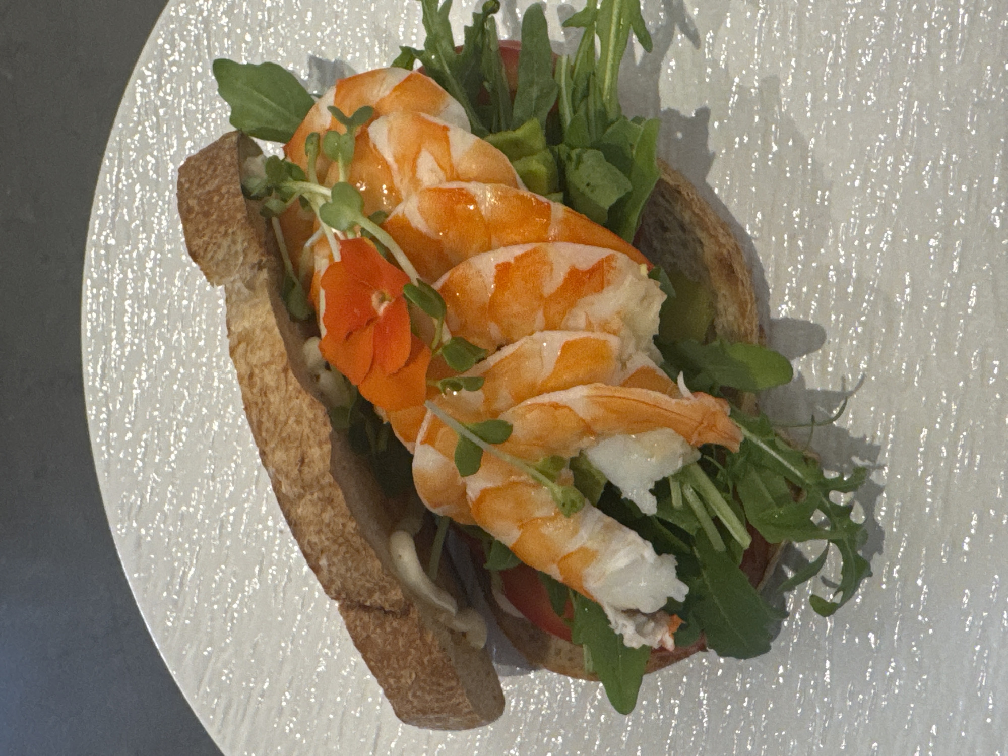 A close-up, top-down shot shows a gourmet shrimp sandwich on a white, textured plate. The sandwich is made with crusty bread, filled with pink shrimp, green arugula, and a few microgreens, and garnished with a small orange flower. The sandwich is artfully arranged, with the shrimp prominently displayed. The background is dark and out of focus, suggesting an indoor setting. The image was taken in Nha Trang, Vietnam, possibly at a restaurant. There are no people visible in the image.