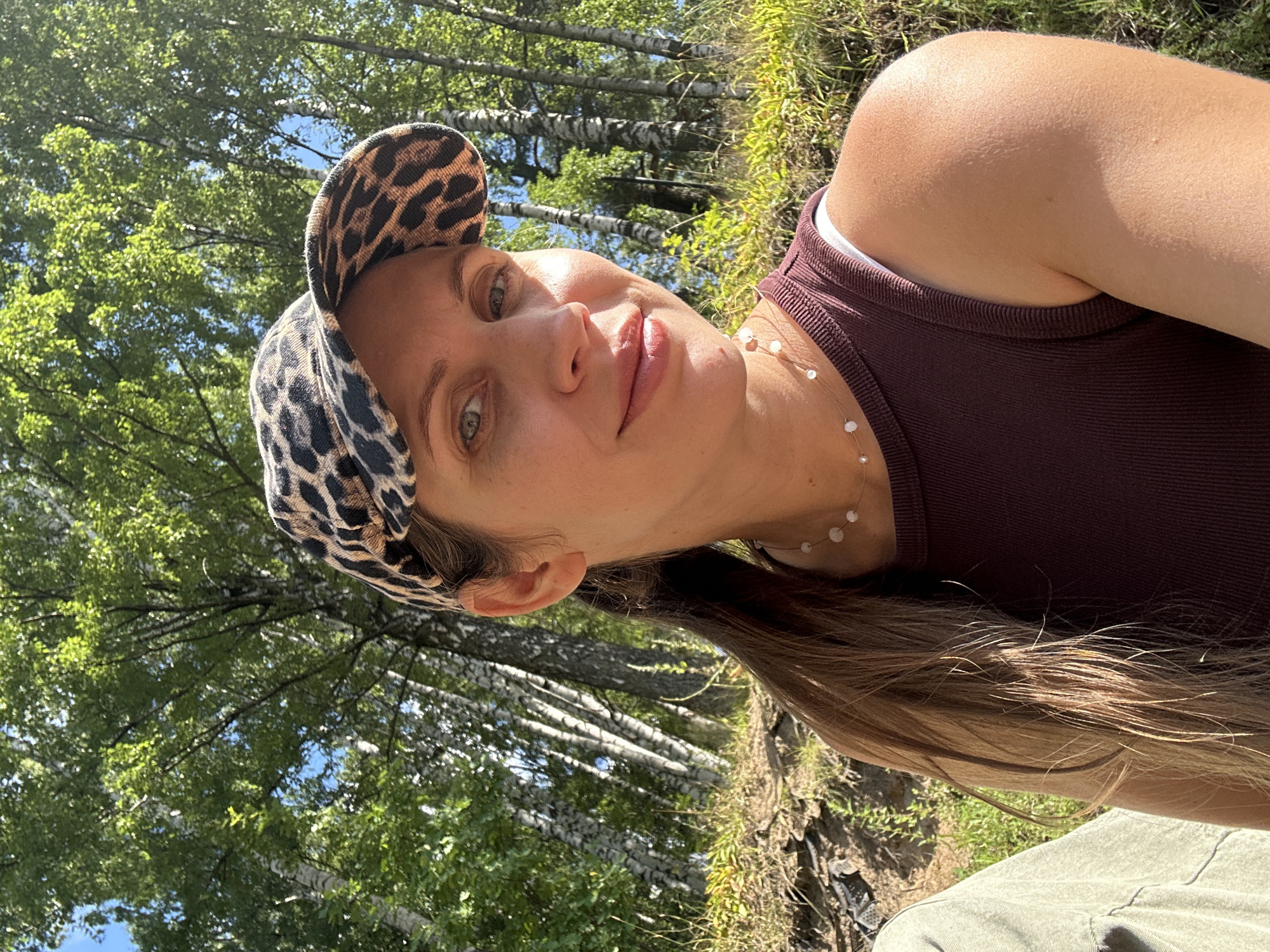 In Vsevolozhsk, Russia, a woman with light brown hair tied in a ponytail wears a leopard-print baseball cap and a maroon ribbed tank top. She has a necklace with small pearls. The woman is looking towards the camera, smiling slightly. She is positioned in front of a backdrop of birch trees with green foliage under a bright sky, indicating an outdoor setting. The lighting suggests it is daytime, possibly late morning or early afternoon. The ground in front of her is covered with dry grass and some dirt. To her left, a portion of another person's clothing, possibly a light green jacket, is visible.
