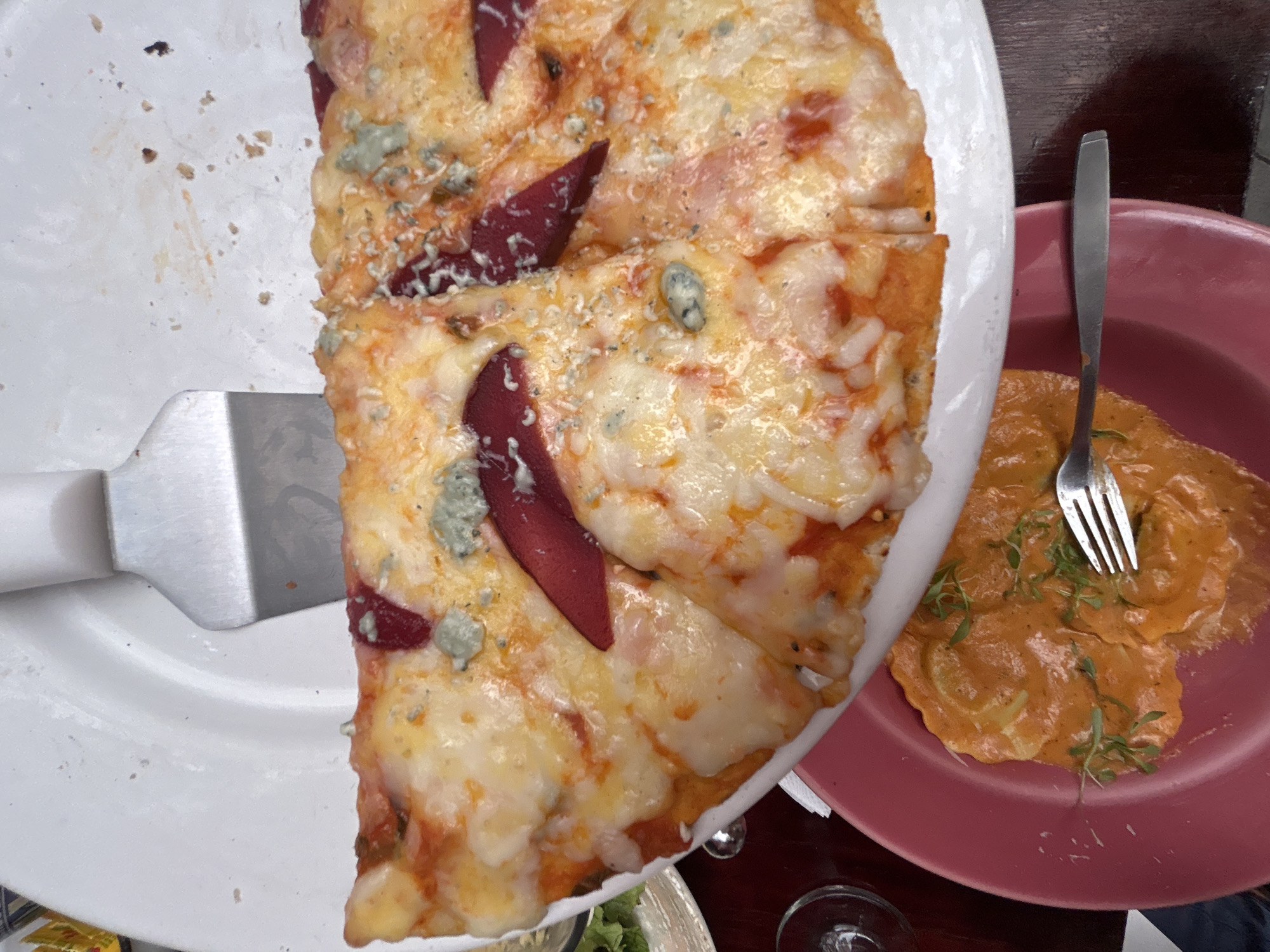 The image shows a close-up of two dishes on a table. On the left, a white plate holds a partially eaten pizza with slices arranged in a semi-circle. The pizza has a light-colored crust and is topped with cheese, tomato sauce, and what appear to be sliced pears and blue cheese. A metal spatula is positioned on the left side of the plate, presumably used for serving the pizza. To the right of the pizza plate is a pink plate containing pasta, likely ravioli, covered in a reddish-orange sauce. A metal fork rests on the pasta. The setting appears to be a restaurant or dining area. The location is unknown.