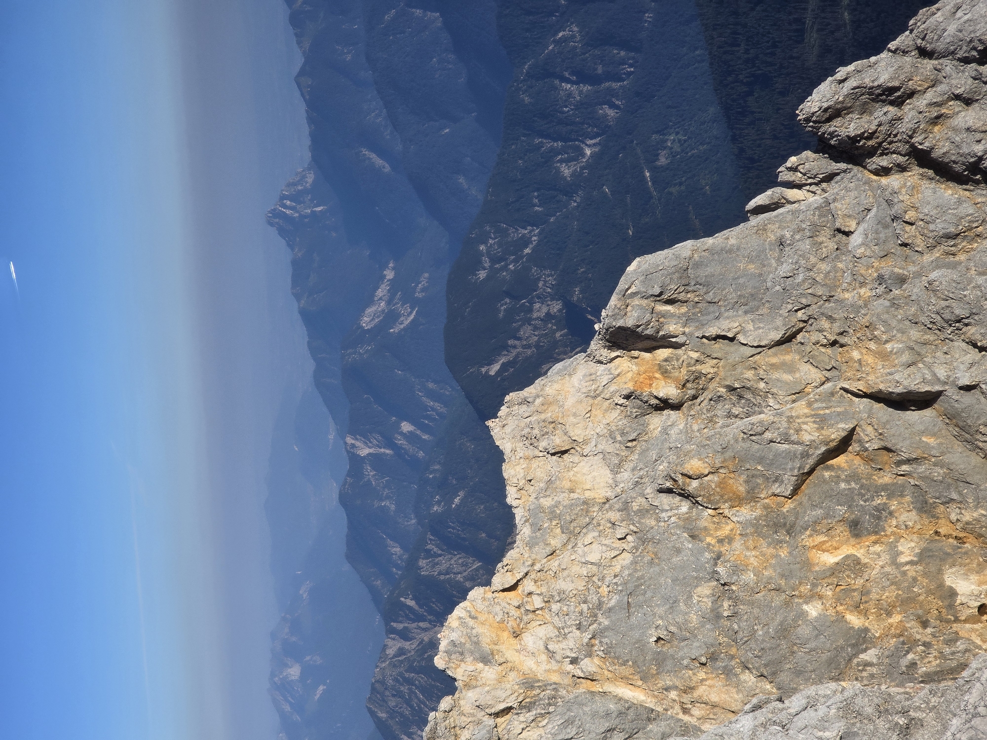 The image depicts a panoramic view of the Alps from a high vantage point, likely on or near the Zugspitze, Germany's highest peak, which is located in the municipality of Garmisch-Partenkirchen. In the foreground, a rugged, sun-drenched rock face dominates the lower left and center of the frame, its texture revealing layers of stone with hues of gray, tan, and orange. Beyond this prominent rock formation, a vast expanse of mountain ranges stretches into the distance.
These mountain ranges are layered in receding shades of blue and purple, indicating atmospheric perspective and the vastness of the terrain. The peaks display a variety of shapes, with some appearing sharp and jagged, while others are more rounded and forested. The lower slopes of some of the closer mountains are covered in dense, dark green forests. The sky above is a clear, bright blue, with a faint haze near the horizon, suggesting it might be a warm, clear day. High in the sky, a thin white contrail from an airplane is visible against the blue. The overall impression is one of immense natural beauty and a sense of elevation and scale. There are no people or man-made objects other than the distant contrail.