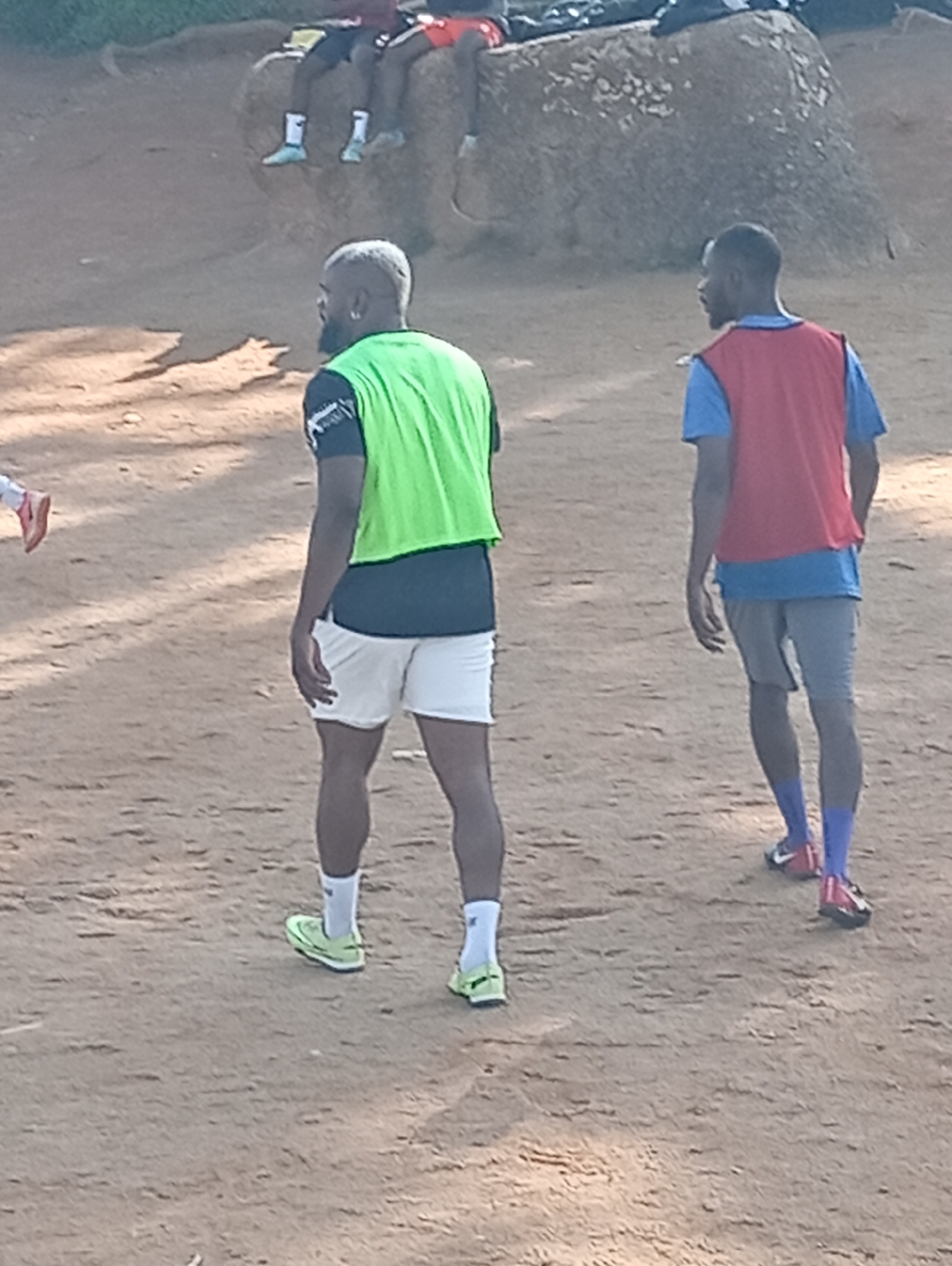 Two young men in athletic attire are standing on a dirt field. The man on the left wears a dark shirt with a bright green vest over it and white shorts. He has a goatee and is looking to his left. The man on the right wears a blue shirt with a red vest over it and grey shorts. He has a short beard and is looking to his right. Both men are wearing athletic shoes and socks. In the background, a large rock is visible, and several other people are seated or standing further back. The environment appears to be an outdoor training or sports ground. This scene is consistent with outdoor recreational activities that might occur in a park or dedicated sports area in Jos, Nigeria.