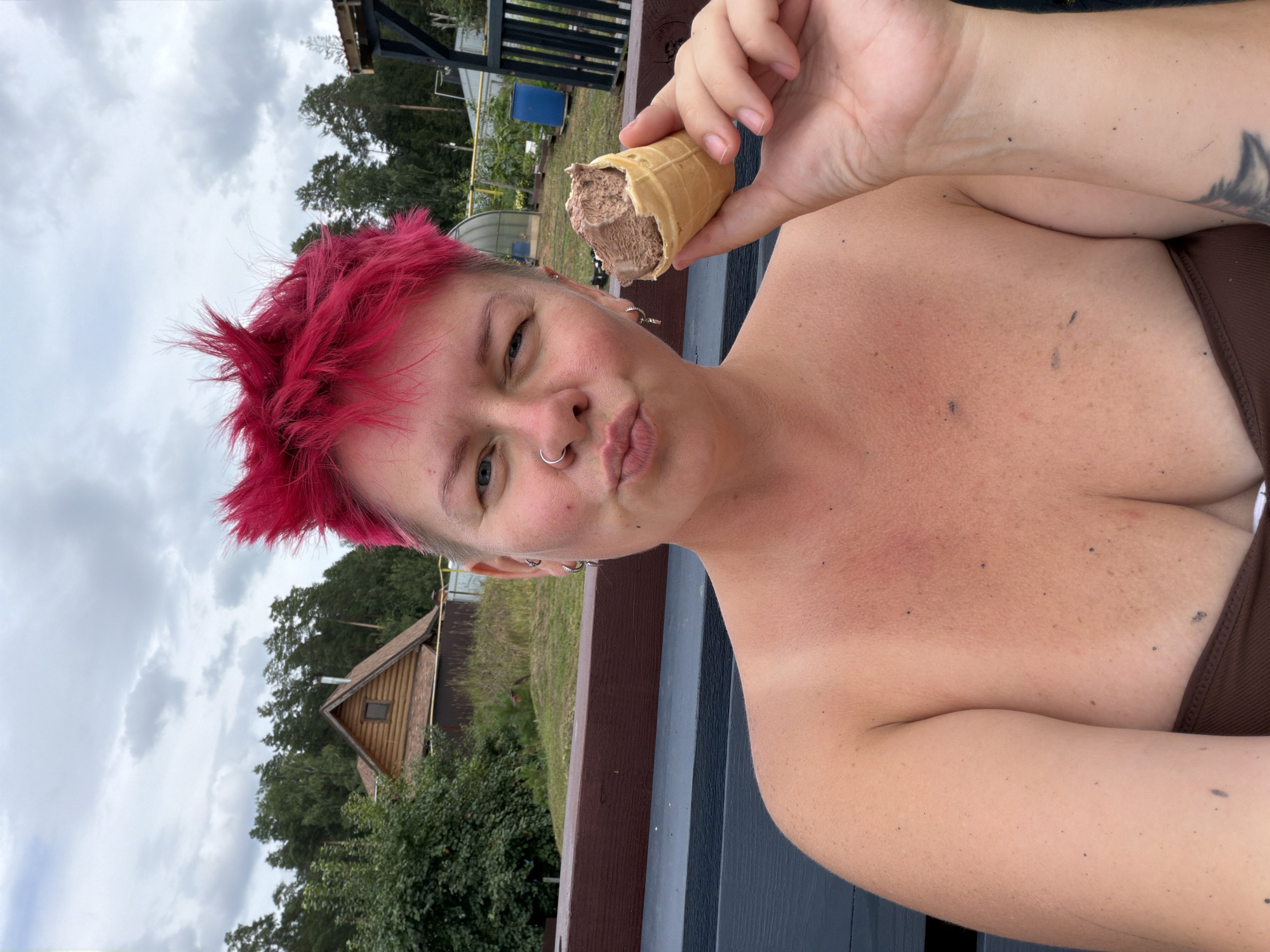 A woman with bright pink, spiky hair and a nose ring is holding a half-eaten ice cream cone in her right hand and making a puckered face at the camera. She is outdoors, appearing to be in a rural or semi-rural setting with a wooden fence in the foreground, a wooden house and trees in the background, and a cloudy sky overhead. The location is identified as Chaikovsky, Russia. The woman is wearing a brown top.
