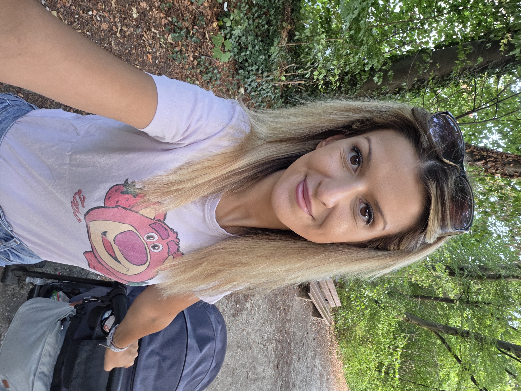 The image shows a woman with blonde hair and sunglasses on her head, taking a selfie while standing next to a stroller in a park. She is wearing a light pink t-shirt with a cartoon character print and blue denim shorts. The park appears to be a wooded area with trees, foliage, and a dirt path. A wooden bench is visible in the background. The stroller is dark grey. The overall lighting suggests it is daytime, likely late afternoon or early evening, given the soft light and shadows. There is no specific indication of the location being in Munich, Germany.