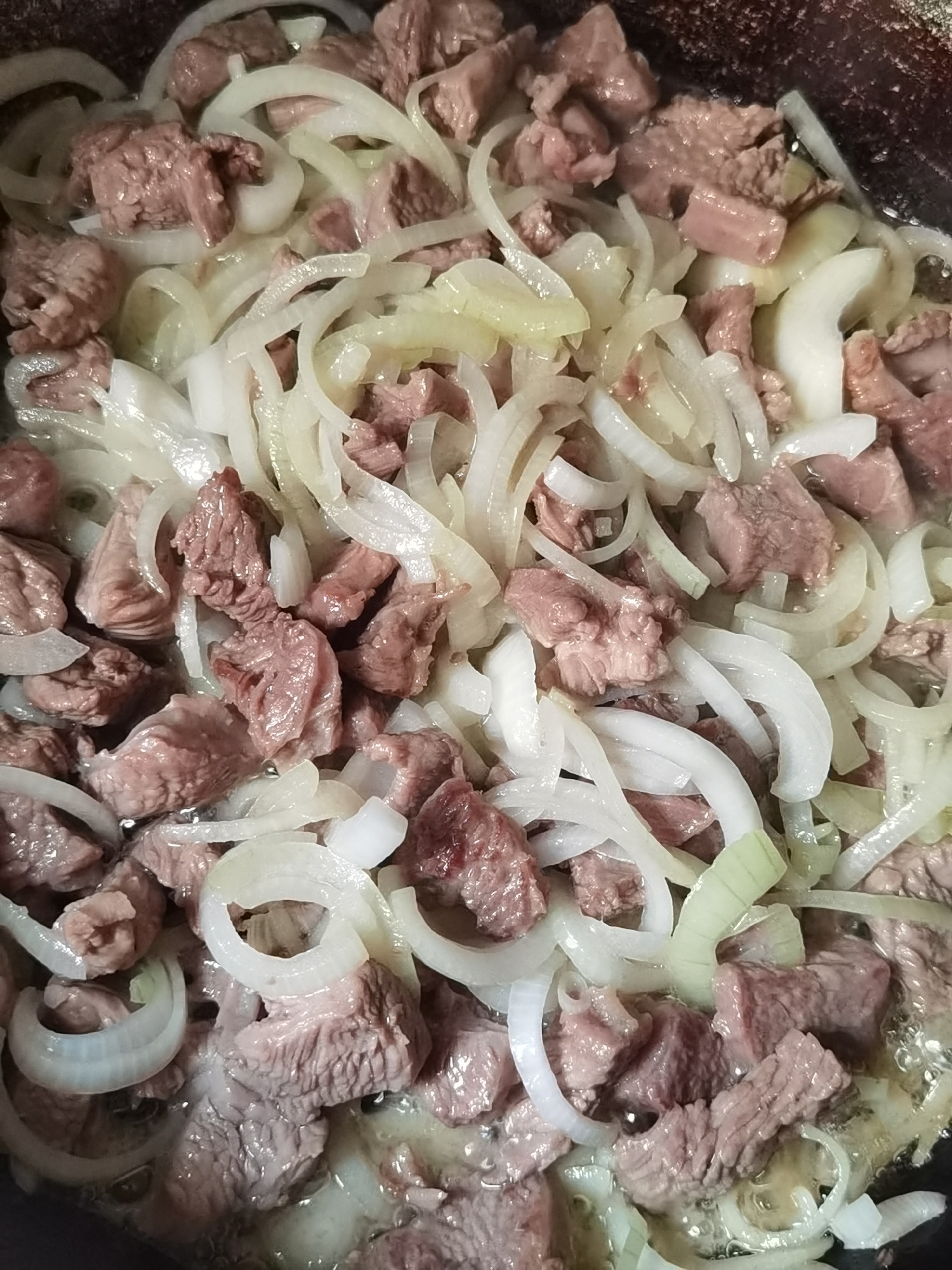 This is a close-up, overhead shot of raw beef and sliced onions being cooked in a pan. The beef is cut into small, irregular chunks and appears to be a deep pinkish-red color. The onions are thinly sliced into rings and have a translucent white to light yellow hue. The ingredients are mixed together and appear to be simmering in some liquid, likely oil or broth, which is visible in small pools and bubbles. The lighting is bright and focused on the food. The background of the pan is dark and slightly textured. This image depicts food preparation, likely in a kitchen environment. There is no discernible location within the city of Chaikovsky, Russia, or any other specific geographical context provided by the visual information.