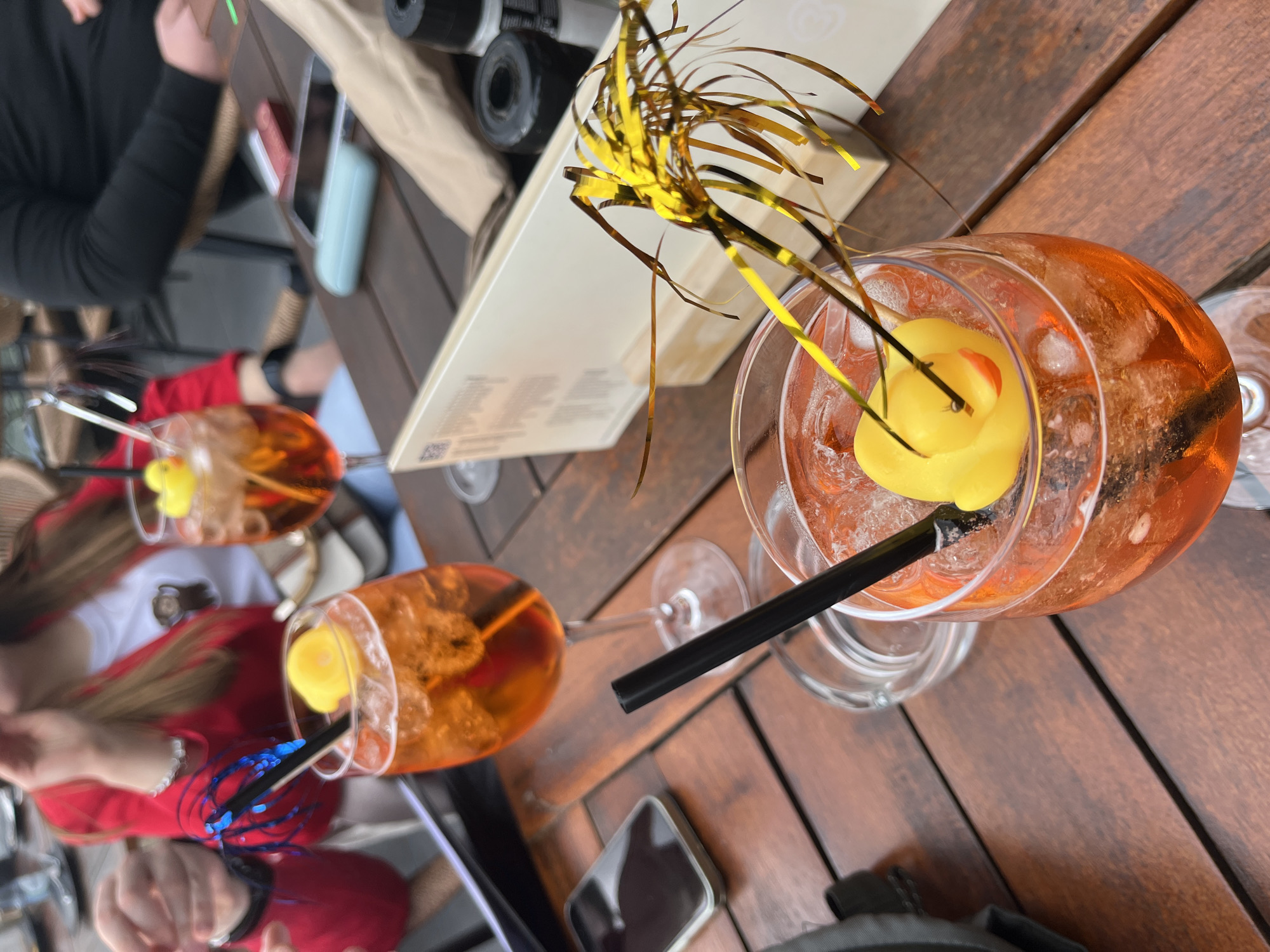 The image shows three Aperol spritz cocktails on a wooden table in Cochem, Germany. Each cocktail is served in a large wine glass with ice and a yellow rubber duck floating on top. Two of the cocktails have decorative metallic accents: one has gold streamers, while the other has blue. Two people are partially visible around the table: one wearing a black shirt and another wearing a red and white shirt. Also on the table are a menu, a cell phone, and some camera equipment.