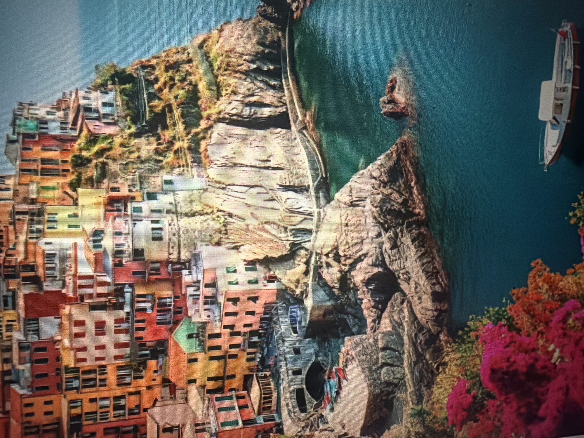 The image depicts a picturesque coastal scene featuring a cluster of colorful buildings perched on a rocky cliffside overlooking a turquoise body of water. The architecture is dense, with buildings of varying heights and hues – including reds, oranges, yellows, and greens – packed closely together. Terraced areas and pathways are visible along the cliff face, leading down to the water. In the foreground, rocks jut out into the sea, and some foliage with bright pink and orange flowers is present. A small white boat with a red hull is gently floating on the water in the lower right portion of the image. The style of architecture and the coastal environment do not correspond to Baan Mae Nam, Thailand, which is known for its fishing village atmosphere and proximity to the Chao Phraya River, not dramatic cliffside settlements overlooking the sea.