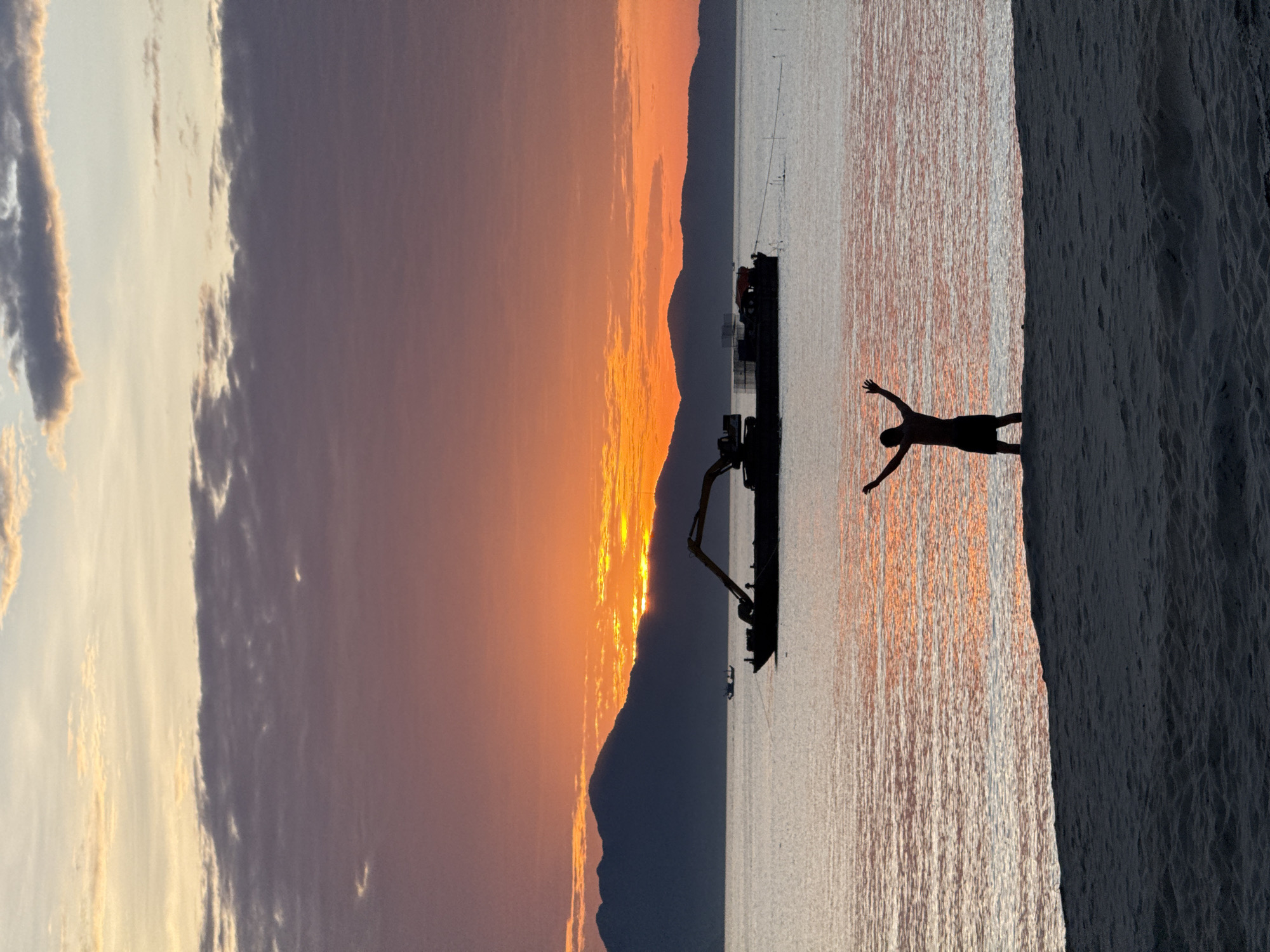 A wide shot captures a sunset scene over a body of water in Đà Nẵng, Vietnam. The sky transitions from light blue and pale yellow at the zenith to vibrant orange and deep red hues closer to the horizon, with scattered dark grey and purple clouds. A range of dark, silhouetted mountains forms the distant background, with the intense orange sun partially obscured behind them. The water reflects the sunset colors, showing streaks of orange and pink, and appears relatively calm. In the mid-ground, a large, dark silhouette of a barge or dredger with an excavator arm extends from its deck, positioned on the water. A smaller, indistinct boat is visible further in the distance to the left of the main vessel. In the foreground, a person stands silhouetted on a sandy beach at the water's edge, facing the sunset with both arms raised high and wide. The sandy beach occupies the bottom portion of the frame, showing dark, granular texture.