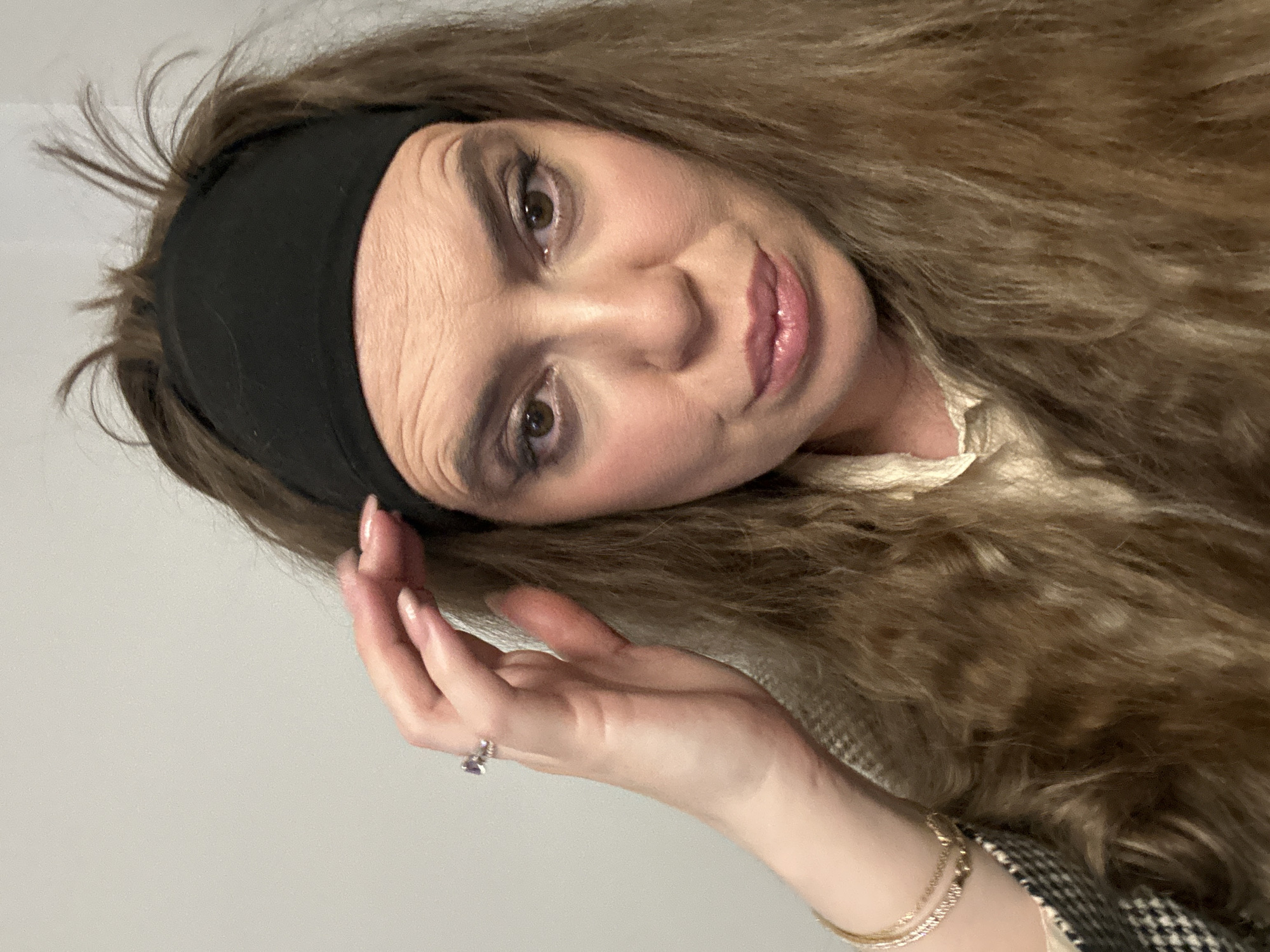 A woman with long, curly, light brown hair is facing the camera. She wears a black headband. Her skin tone is fair, and she is wearing makeup, including eye shadow and lipstick. She has brown eyes. Her right hand is near her face, with fingers slightly spread. She wears a ring on her ring finger and gold bracelets on her wrist. She is wearing a light-colored top with a textured jacket, suggesting a houndstooth pattern. The background appears to be a plain, light-colored wall. There is no indication of Tbilisi, Georgia in the image. The location cannot be ascertained.