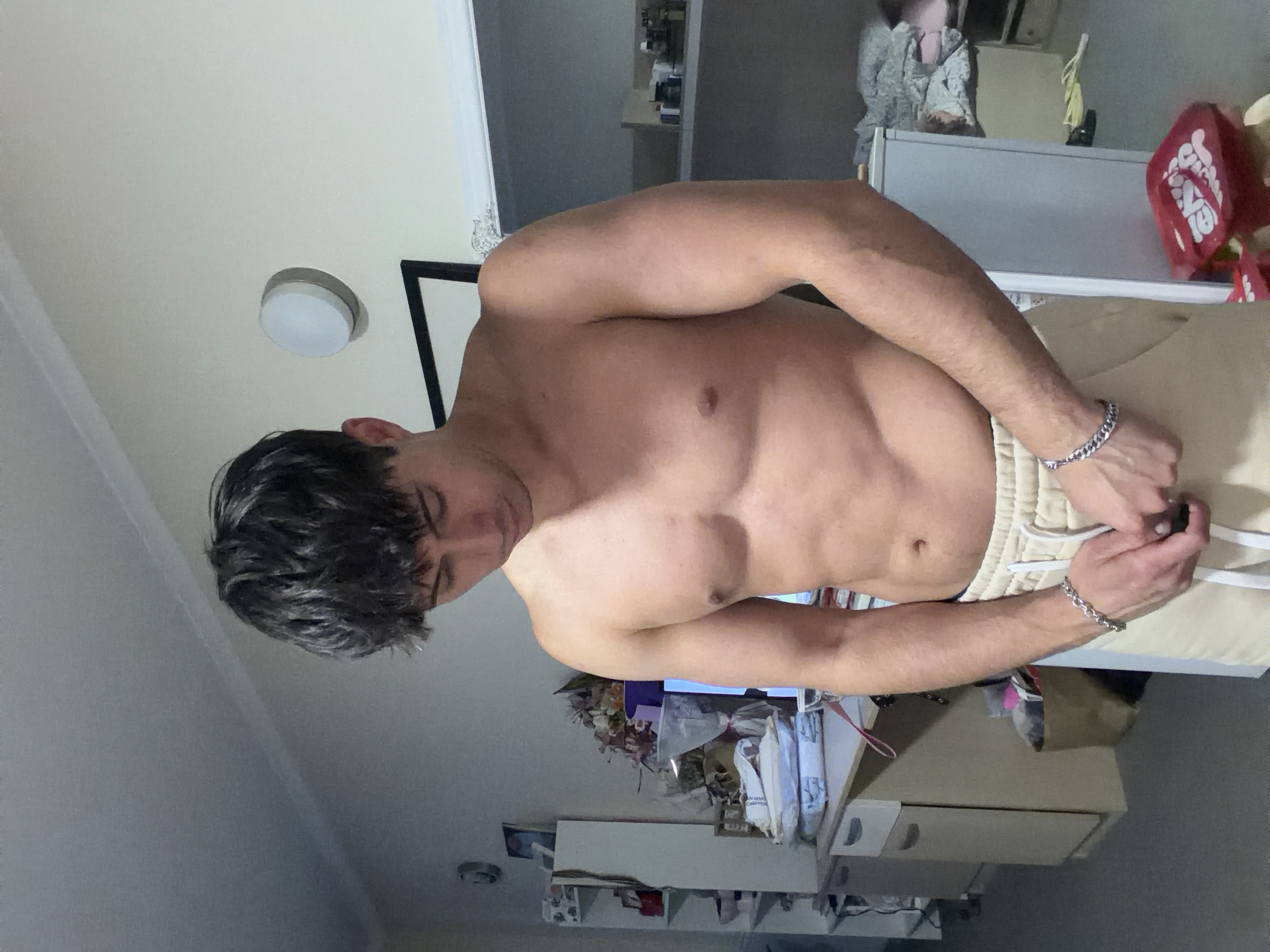 The image, rotated 90 degrees clockwise, captures a shirtless young man in an indoor setting, possibly in Sofia, Bulgaria. The man has short, dark, messy hair and is looking downwards. He exhibits a lean, muscular physique and is wearing light-colored shorts or sweatpants. Silver chain bracelets are visible on both of his wrists, and his hands are actively engaged in adjusting or tying the drawstring of his shorts.
The background shows a room with light-colored walls and ceiling, featuring a circular flush-mount light fixture. A black-framed mirror or picture frame is partially visible behind the man's head and shoulder. Further in the background, there is a white or light-colored dresser or cabinet, cluttered with various items, including what appears to be a doll or figure on top and a red object with indistinct, unreadable white text. The overall impression is a lived-in room with some clutter. The lighting suggests artificial indoor illumination. No specific time of day or outdoor weather conditions are discernible.