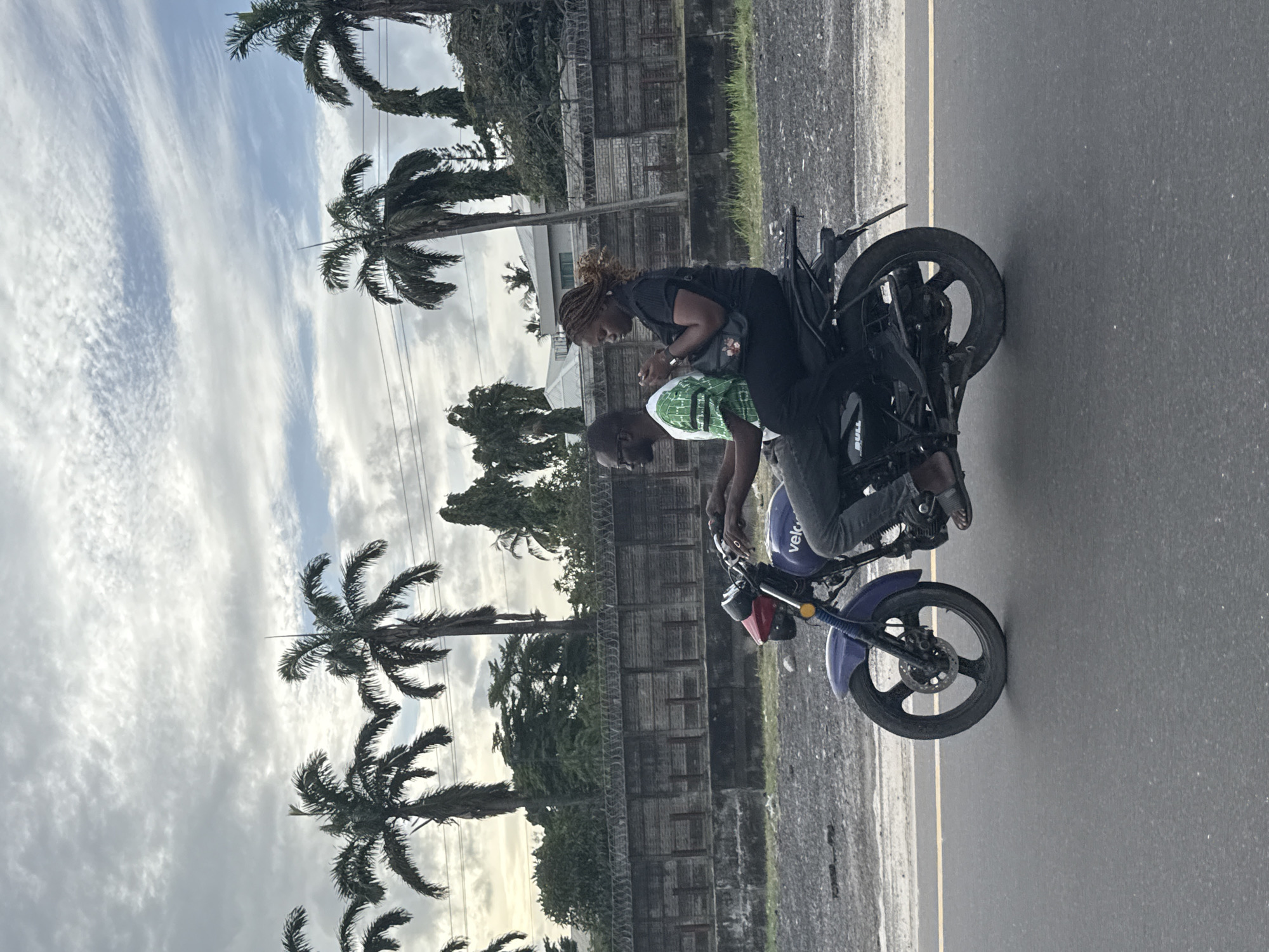 The image captures two individuals on a motorbike by the side of a road in Ojomu, Nigeria, under an overcast sky.
In the foreground, a blue or purple motorbike is parked or moving slowly on asphalt. A person, appearing male, is seated in the front, wearing a green and white patterned shirt. Behind them, a second person, appearing female, is riding pillion. She is dressed in dark clothing and has braided hair, leaning slightly forward and looking down, possibly at a phone or an object in her hands, or engaging with the driver. Visible text on the motorbike includes "velo" on the front and "BULL" near the rear wheel.
The background is dominated by several tall palm trees, suggesting a tropical or subtropical environment. Behind the trees and the subjects, a long, grey concrete wall topped with barbed wire runs horizontally across the frame, indicating a secured boundary. Overhead power lines are faintly visible against the sky. The sky is mostly covered with clouds, exhibiting varied patterns from wispy cirrus-like streaks to denser, more uniform cloud cover, suggesting a daytime scene with diffused lighting. The road is asphalt, with a subtle yellow line visible along one edge.
