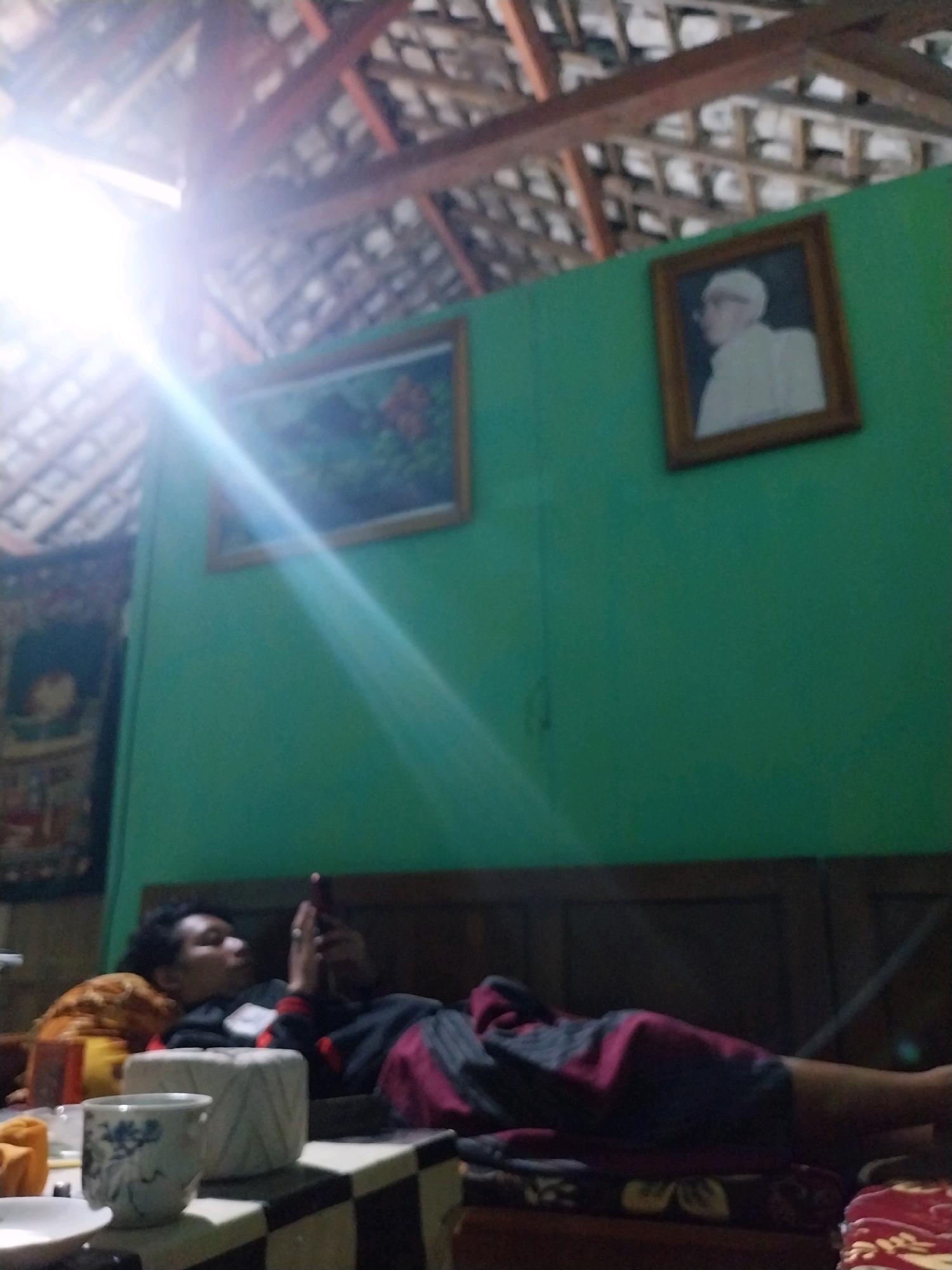 The image depicts an indoor scene, possibly a bedroom, with a thatched roof and exposed wooden beams visible overhead. The walls are painted a vibrant green. On the wall hangs a framed portrait of a person wearing a white cap and robe. Another framed picture, showing what appears to be flowers, is also on the wall, partially obscured by a bright light flare.
In the foreground, a person is lying on a bed, looking at a smartphone. The person is wearing a dark jacket with red accents and striped pants. They are covered by a blanket with a patterned design. Beside the bed, on a surface with a checkered pattern, is a white ceramic mug with blue floral designs and other objects, including what appears to be a plate. The lighting in the room is dim, with a strong light source creating a significant flare that streaks across the upper left portion of the image. The overall impression is one of a domestic setting.