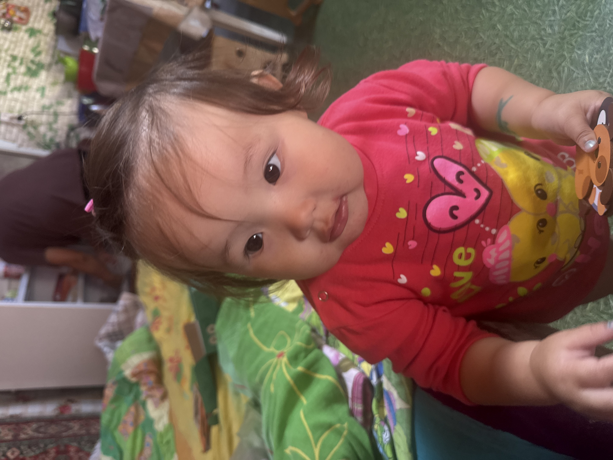 In Yabogan, Russia, a young child with dark hair is seated on a bed covered with colorful blankets. The child is wearing a bright pink long-sleeved shirt adorned with hearts and cartoon characters. Their right hand is holding a small, brown, animal-shaped toy. The background suggests an indoor setting with shelving units and a person partially visible in the distance. The scene is illuminated by natural light, creating a warm and intimate atmosphere.