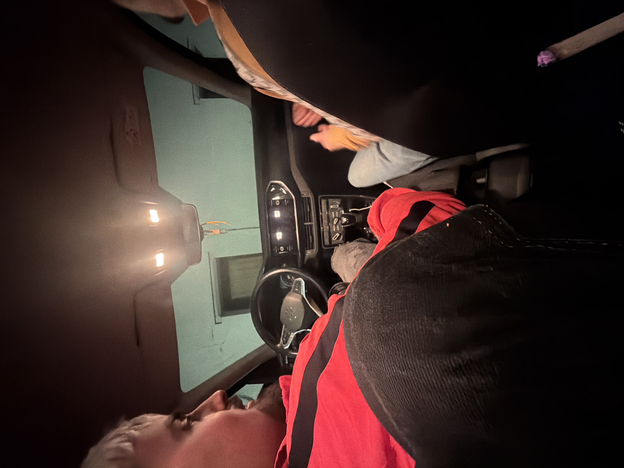 A rotated image shows a person lying in the driver's seat of a car. The person is facing toward the top of the image. The car's interior is visible, including the steering wheel, dashboard, and center console. Another person is in the passenger seat. The location cannot be determined from the image alone.