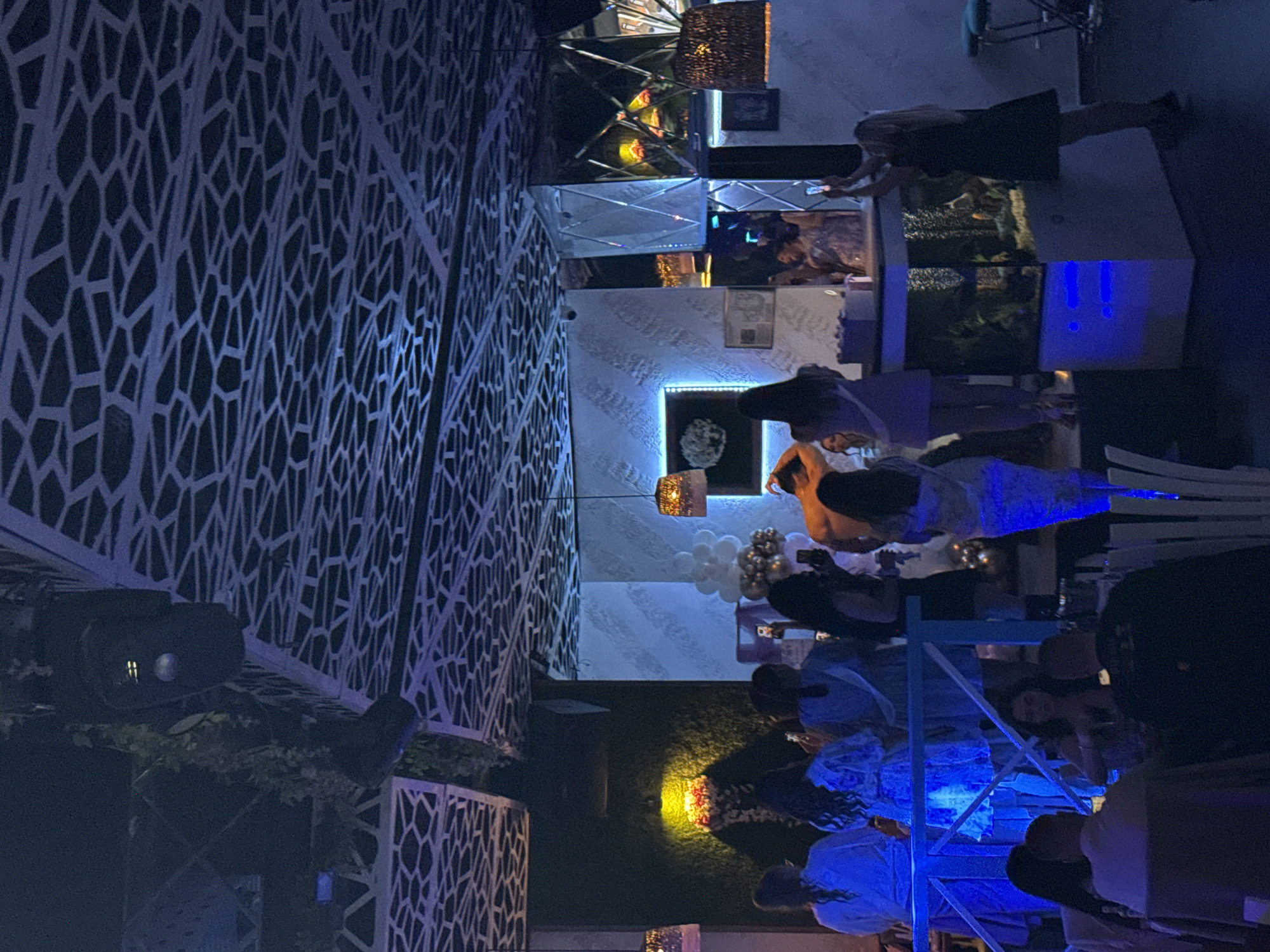 The image shows an outdoor evening event with people gathered at tables and near what appears to be a bar area. The lighting is low, with various light sources contributing to a predominantly blue and yellow cast. Several individuals are visible, some standing and others seated, engaged in conversation or interaction. The environment includes decorative lattice structures and a lighting display. The event likely takes place at the Vogue Club, in the city of Plovdiv, Bulgaria.