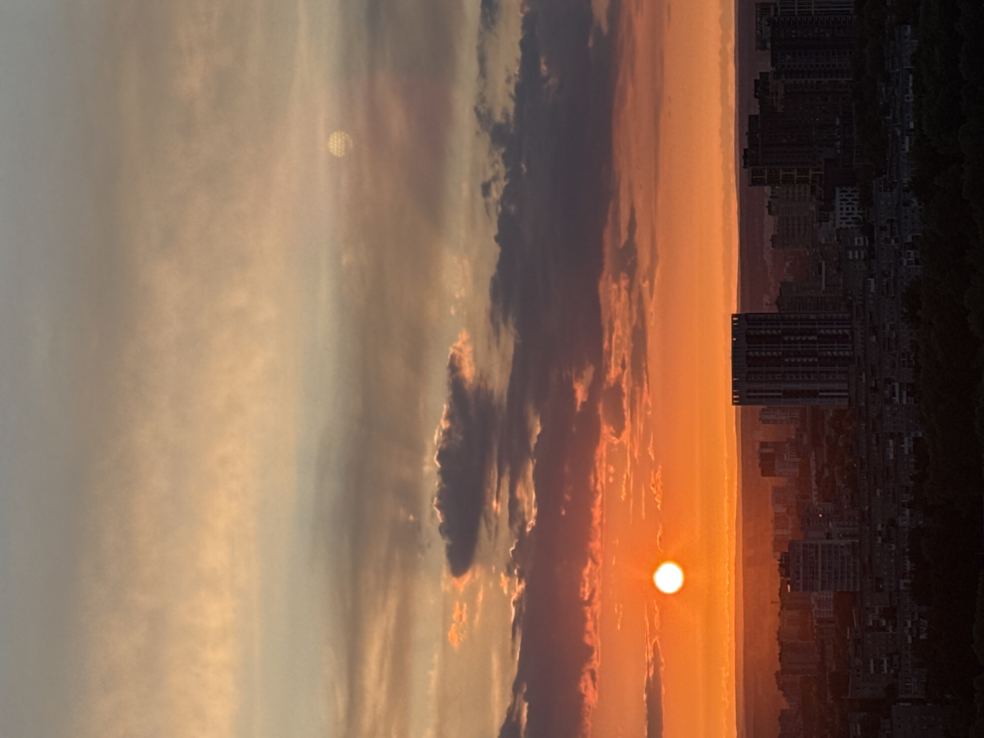 The image captures a panoramic view of an urban landscape at either sunrise or sunset, centered on the vibrant sky and silhouetted city. The sky is dominated by warm, intense orange and red hues near the horizon, gradually transitioning to softer golden, grey, and light blue tones higher up and to the left.
A brilliant, round, and intensely bright sun is prominently featured near the right side of the horizon, casting a strong glow across the lower sky and illuminating the edges of nearby clouds. Various cloud formations are scattered across the sky; some are dark and voluminous, creating dramatic contrasts, while others are more wispy and diffuse, catching the golden light. In the upper left quadrant, a fainter, hazy circular light source, possibly the moon or the sun obscured by denser clouds, is visible, exhibiting a textured appearance.
Below the colorful sky, a sprawling urban skyline is silhouetted in dark tones, stretching across the lower portion of the image. Numerous buildings, including multi-story structures, appear as dark masses against the bright background. A faint line of distant landforms, possibly hills or mountains, is visible along the horizon behind the city. The overall atmosphere conveys a sense of serene grandeur, depicting the meeting of nature's spectacle with human habitation. The scene is set in Сокол, Russia. No visible text is present in the image.