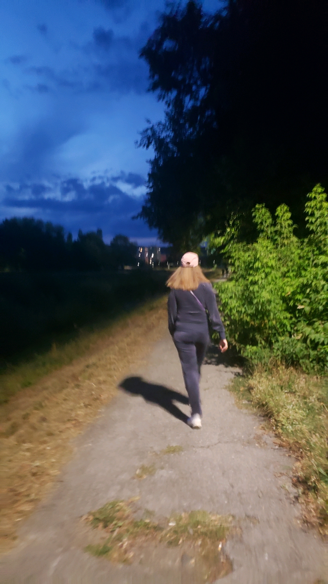 An individual walks away from the camera down a paved path in the twilight. The individual is wearing a dark tracksuit, a pink baseball cap, and white shoes. They are casting a shadow on the path. To the right of the path, there is dense green foliage, and to the left, the ground is covered in dry grass. In the background, a dark silhouette of trees borders a distant cityscape with visible lights. The sky above is a deep blue with some cloud formations. The scene appears to be an outdoor path in a park or along a waterfront within the city of Prešov, Slovakia, likely during the evening hours.