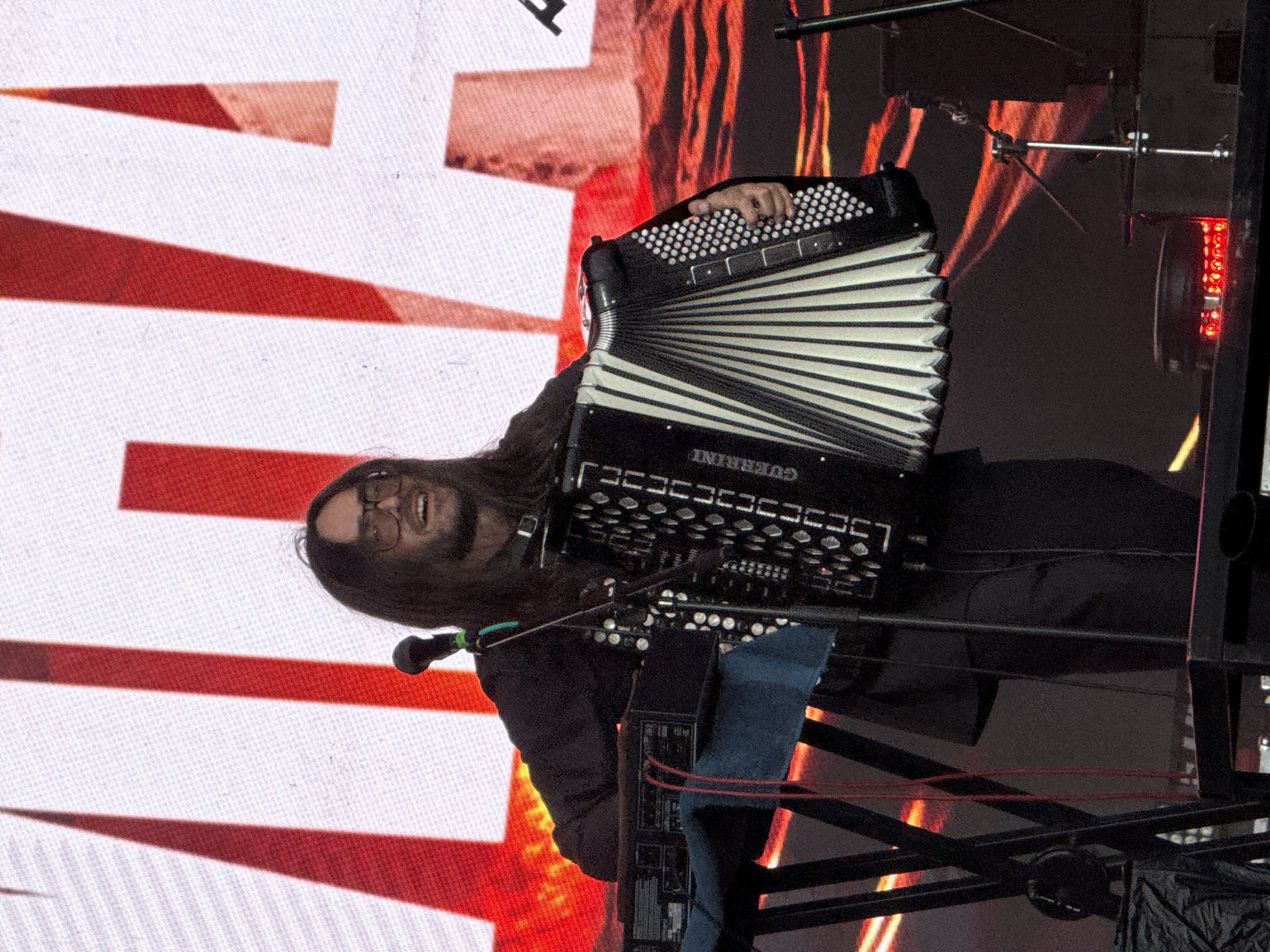 A man with dark, shoulder-length hair and glasses is playing an accordion on a stage. He is wearing a dark jacket and dark pants. He is smiling and looking to his right. The accordion is black and white with silver buttons. To his left, there is a keyboard on a stand with red wires. In the background, there is a large screen with red and white diagonal stripes. The stage appears to be outdoors, as indicated by the lighting and the general atmosphere. There is no discernible information in the image to suggest a specific location within Lagunovo, Russia.