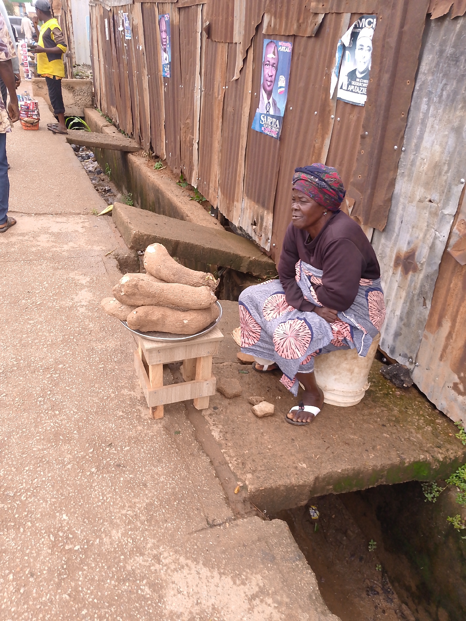 The image captures a street scene in Bukuru, Nigeria, showing a woman selling yams from a roadside stall. An elderly woman, the central figure, is seated on an inverted white plastic bucket on a concrete pavement. She wears a dark long-sleeved top, a vibrant patterned wrapper (skirt) with blue and pink designs, and a multi-coloured headscarf. Her expression is calm, appearing contemplative or waiting.
In front of her, on a small, light-coloured wooden stool, rests a metal platter holding a pile of large, light brown yams. Behind the woman, a rusty corrugated iron wall forms the backdrop, typical of informal structures. Several political posters are affixed to this wall. One blue poster features a man's portrait and the text "SARPITA ANDO DON" along with "SA". Another black and white poster displays a different man's portrait and includes text such as "AVICHI", "MANIY", and "HON. (DR) AGULAK WE APUTAZIE".
To the left of the woman, other people are partially visible, including a person in a yellow and black shirt standing near a display of bottled drinks. A concrete gutter or drainage channel runs along the edge of the pavement, with some sections covered by concrete slabs. The ground is a textured, light brown concrete surface. The lighting suggests it is daytime, possibly an overcast day, given the soft, diffused light and lack of strong shadows. The scene depicts a common sight of street vending and daily life in a bustling local environment.
