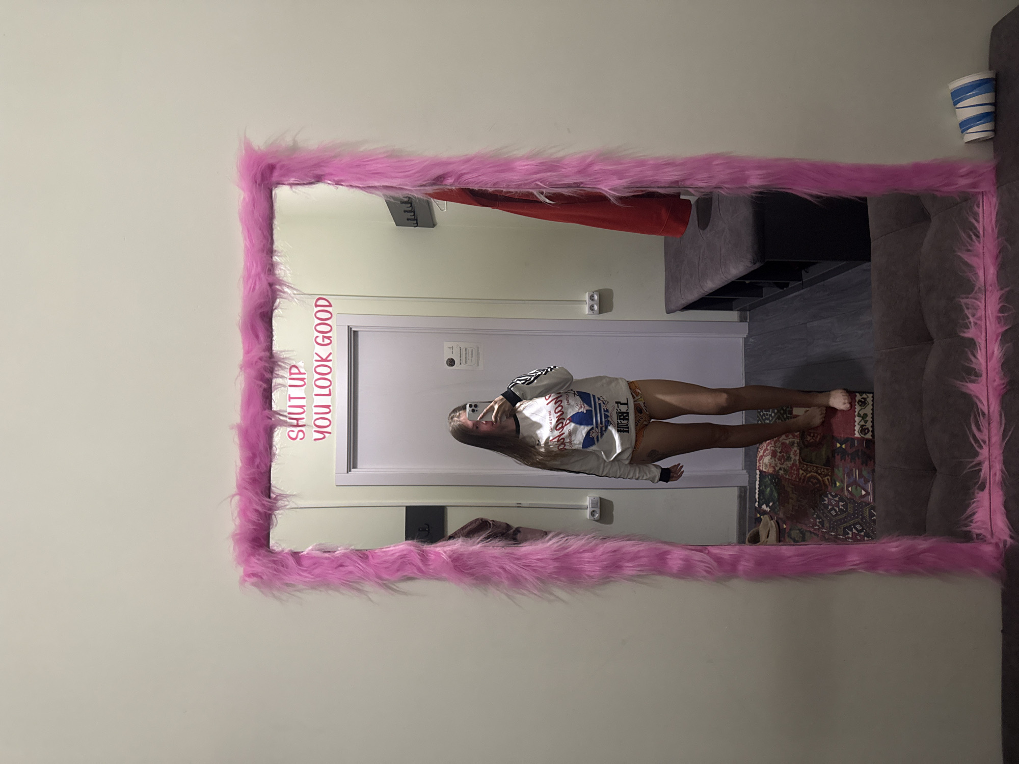 A young woman is taking a selfie in a mirror with a pink, furry frame. The text "SHUT UP YOU LOOK GOOD" is written on the upper left corner of the mirror. The mirror is mounted on a wall, reflecting a room with a doorway, a clothes rack with a red garment, a small table, and a rug. A portion of a sofa is visible on the right side of the mirror. There are blue and white cups on top of the sofa. The location is Batumi, Georgia.
