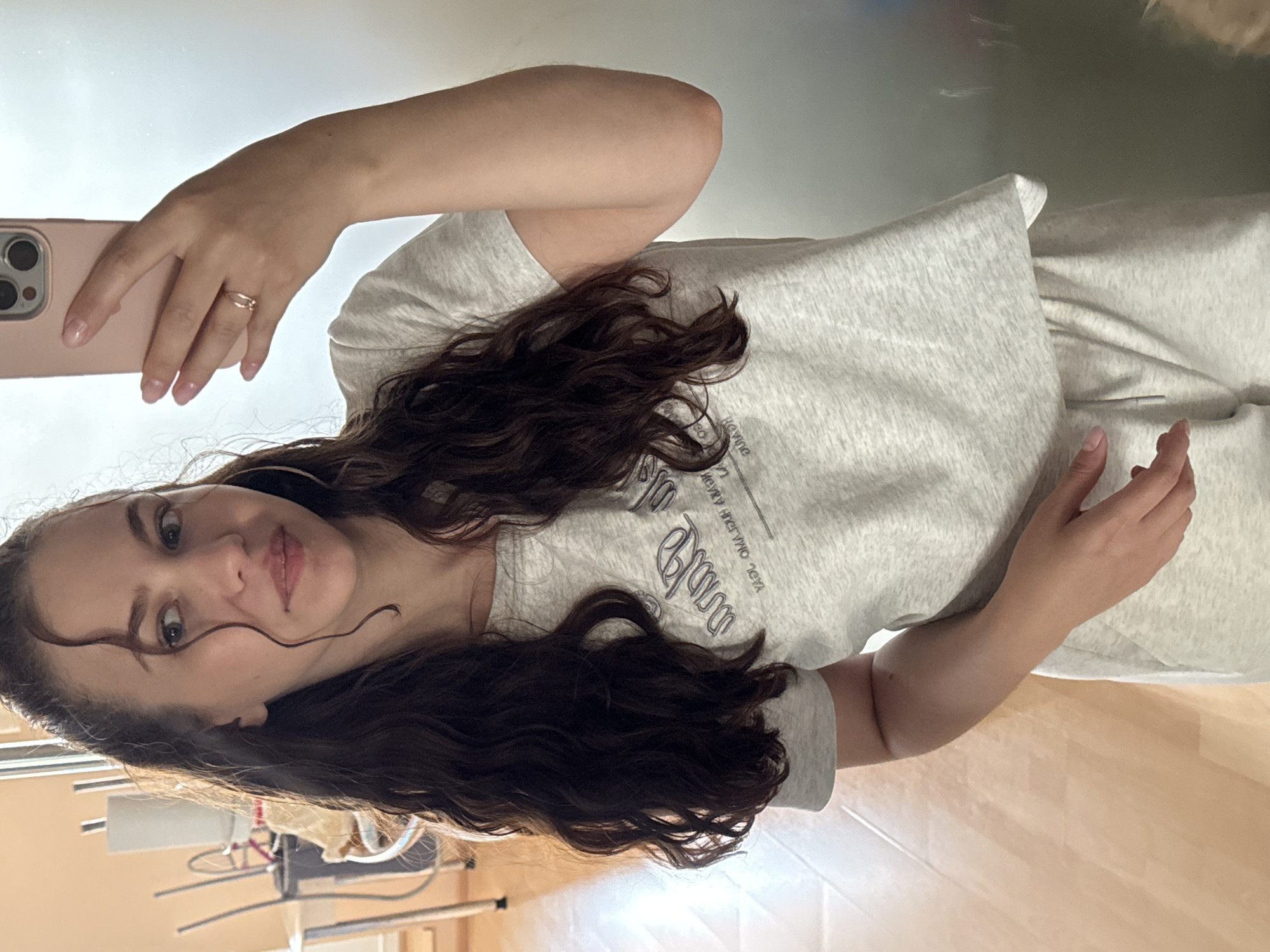 A young woman with long, dark, curly hair is taking a selfie in a room. She is wearing a light gray t-shirt with a dark gray graphic print and light gray sweatpants. She is holding a pink smartphone in her right hand, which is angled to capture her reflection in a mirror. A thin gold ring is visible on her left hand. The background shows a wooden floor and some furniture, including chairs and a waste bin. The overall impression is that of a casual, indoor setting, possibly a home. The image is set in Chaikovsky, Russia.