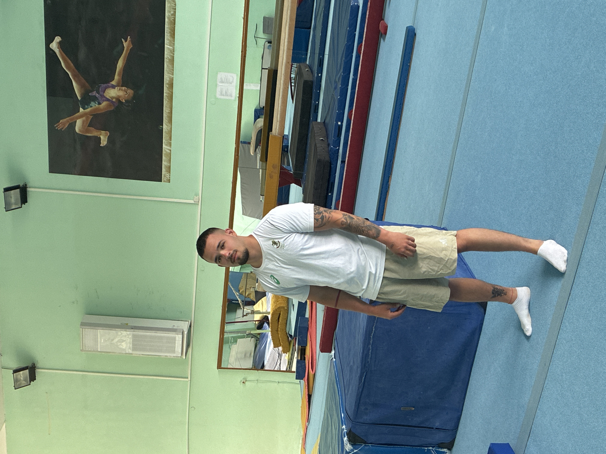 A man in a white t-shirt and khaki shorts stands in a gymnasium. He is wearing white socks. There is a poster of a female gymnast performing on the wall. The gym has blue and light green walls, with mats, balance beams, and a mirror visible. The man has tattoos on his arms and leg. No specific landmark identifies it as Blagoevgrad, Bulgaria.