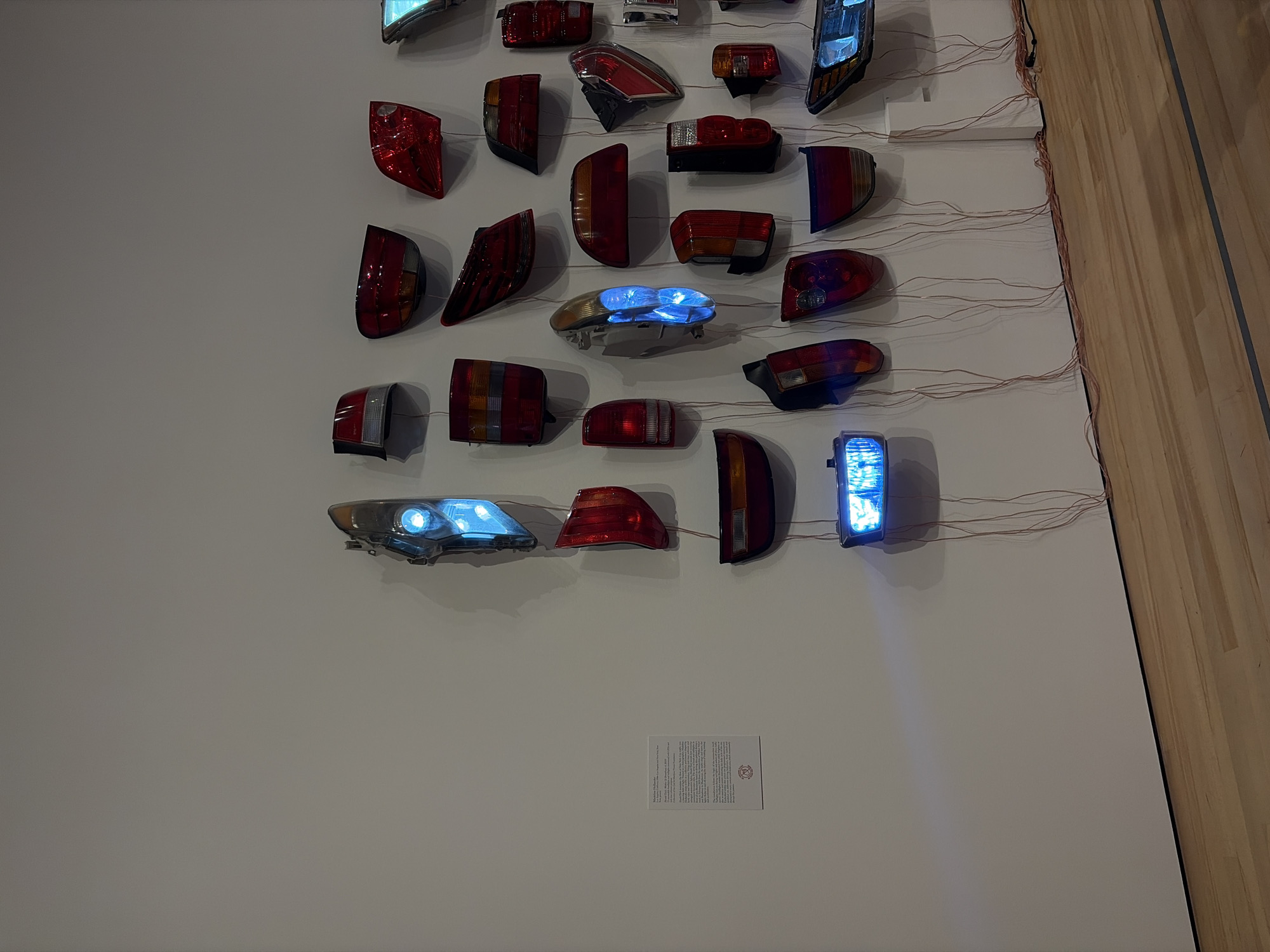 This image shows an art installation on a white wall. The installation features various car taillights and headlights arranged in a grid-like pattern. Some of the lights are illuminated with a blue glow. Thin wires run from each light towards the upper right, likely connecting them to a power source. A small, rectangular informational plaque is visible on the wall below the lights. The right edge of the image shows a wooden wall, suggesting this installation is indoors. This is located in the city of Austin, United States of America.