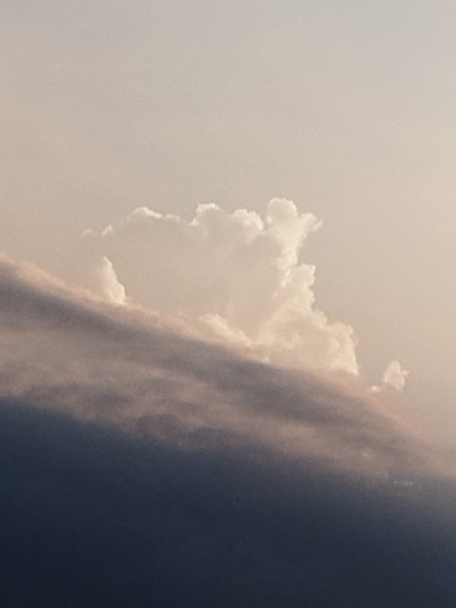 The image shows a sky with clouds. A large, fluffy white cloud is positioned in the upper center, illuminated from the front, suggesting sunlight. Below it, a band of darker, textured clouds runs horizontally across the frame, obscuring the lower portion of the sky. The background sky is a soft, diffused pale color. There are no discernible elements of the city of Vidra, Romania, visible in this image. The scene is entirely focused on the atmospheric conditions, specifically the arrangement and appearance of clouds.