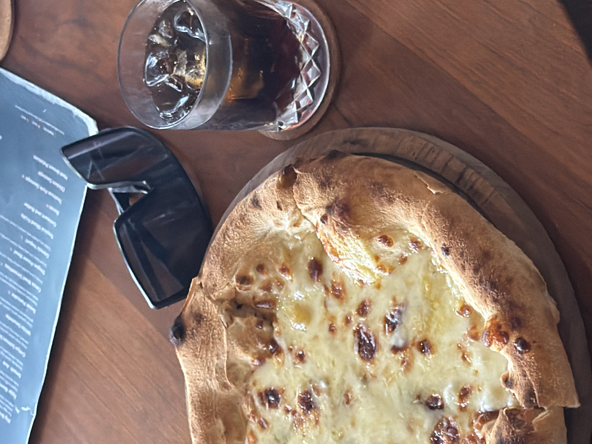 On a wooden table, a pizza sits on a round wooden serving board. To the left are black sunglasses and a menu. A glass of dark liquid with ice rests on a coaster above the pizza. This scene is likely set in Tegal Suci, Indonesia.
