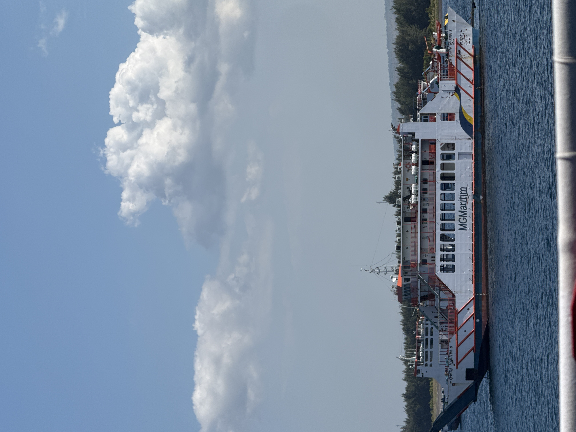A large white and orange ferry, the "GARDA MARITIM 8," sails on a blue body of water under a bright blue sky dotted with fluffy white clouds. The ferry has multiple decks and windows. In the background, a line of green trees forms a treeline along the shore, with a faint outline of hills in the distance. The scene appears to be a sunny day at sea, likely near the coast. The location is identified as the city of Gunung Malang, Indonesia. There are no people visible in the frame.