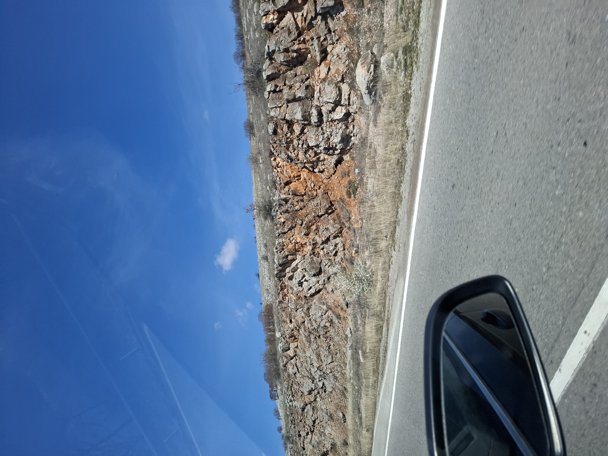 The image depicts a view from a moving vehicle on a road near Bogyovtsi, Bulgaria. The road is asphalt and has a white line marking the edge. To the left of the road is a rocky, barren hillside with sparse vegetation. The sky is clear and blue, with a few small clouds. The side mirror of the car is visible in the lower right corner of the frame.