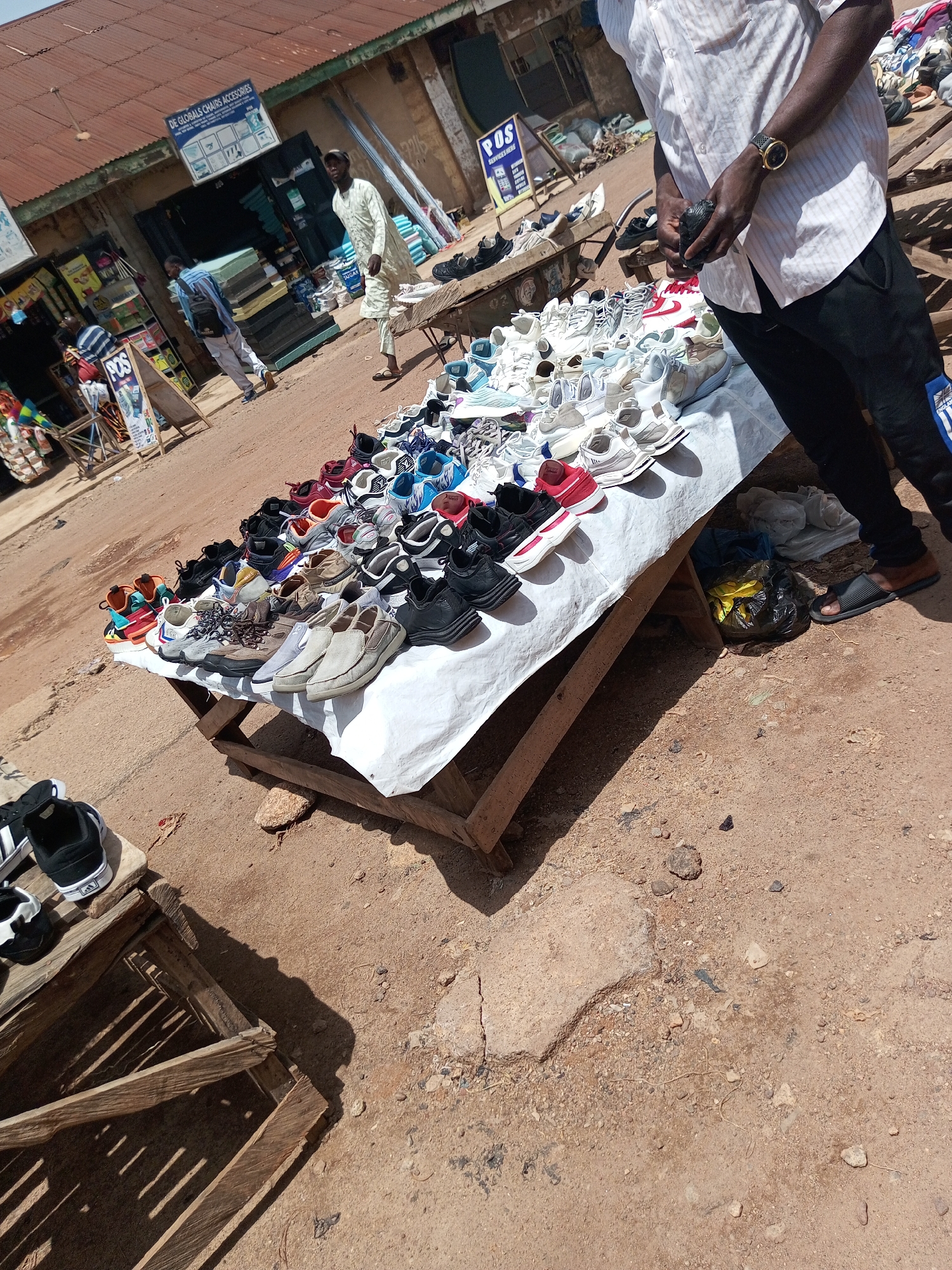 The image depicts an outdoor market scene in Bukuru, Nigeria. In the foreground, a table covered with a white sheet displays a large assortment of sneakers and casual shoes. The shoes are arranged in rows, with various colors and styles visible. To the left of the table, a wooden crate contains a few pairs of black and white sneakers.
In the middle ground, a man in a white patterned shirt and dark pants stands holding a small dark object in his hands. He is wearing a wristwatch on his left wrist. Behind him, a person wearing a light-colored tunic walks away from the camera. Further back, several shops line the street, with awnings and signage indicating businesses. One sign reads "DE GLOBALS CHAIRS ACCESSORIES". The ground is dirt and appears somewhat dusty. The overall environment suggests a bustling commercial area.