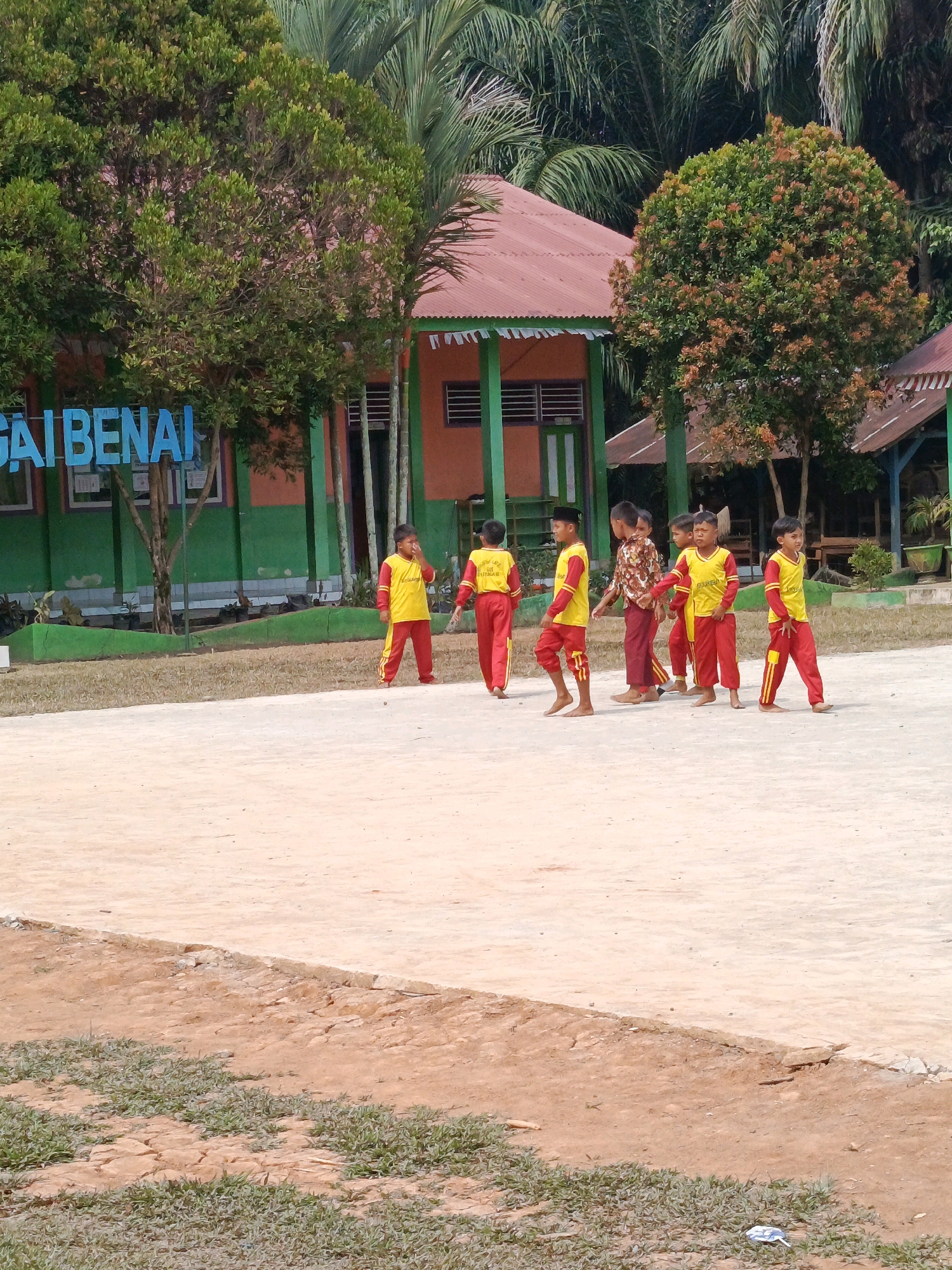 The image depicts an outdoor scene in what appears to be a schoolyard or community area in Megang Sakti IV, Indonesia. In the foreground, a sandy, light-colored surface dominates the frame, with a strip of dry grass and earth bordering the bottom edge.
In the middle ground, a group of seven young boys, all appearing to be of primary school age, are spread across the sandy area. They are mostly dressed in matching yellow shirts with red accents and red pants. Some are standing, while others are walking or in motion. One boy on the far left is facing away from the camera, his back to the viewer, while another boy in the center is looking to his left. The boy third from the right is facing the camera and appears to be wiping his face or nose. Another boy on the far right is facing the camera with his body angled to the left. The interactions between the boys seem casual, with no obvious signs of organized activity or distress.
The background features a low-rise building with a reddish-orange roof and light orange walls, accented with green trim and pillars. Windows with horizontal slats are visible on the building's facade. Several trees with lush green foliage are scattered throughout the scene, some partially obscuring the building. In the far background, dense green foliage suggests a tropical environment. A blue sign with white lettering, partially visible on the left side of the image, bears the text "SAI BENAI," which is likely part of a larger place name or school designation. The overall impression is a sunny day in a rural or semi-rural Indonesian setting.