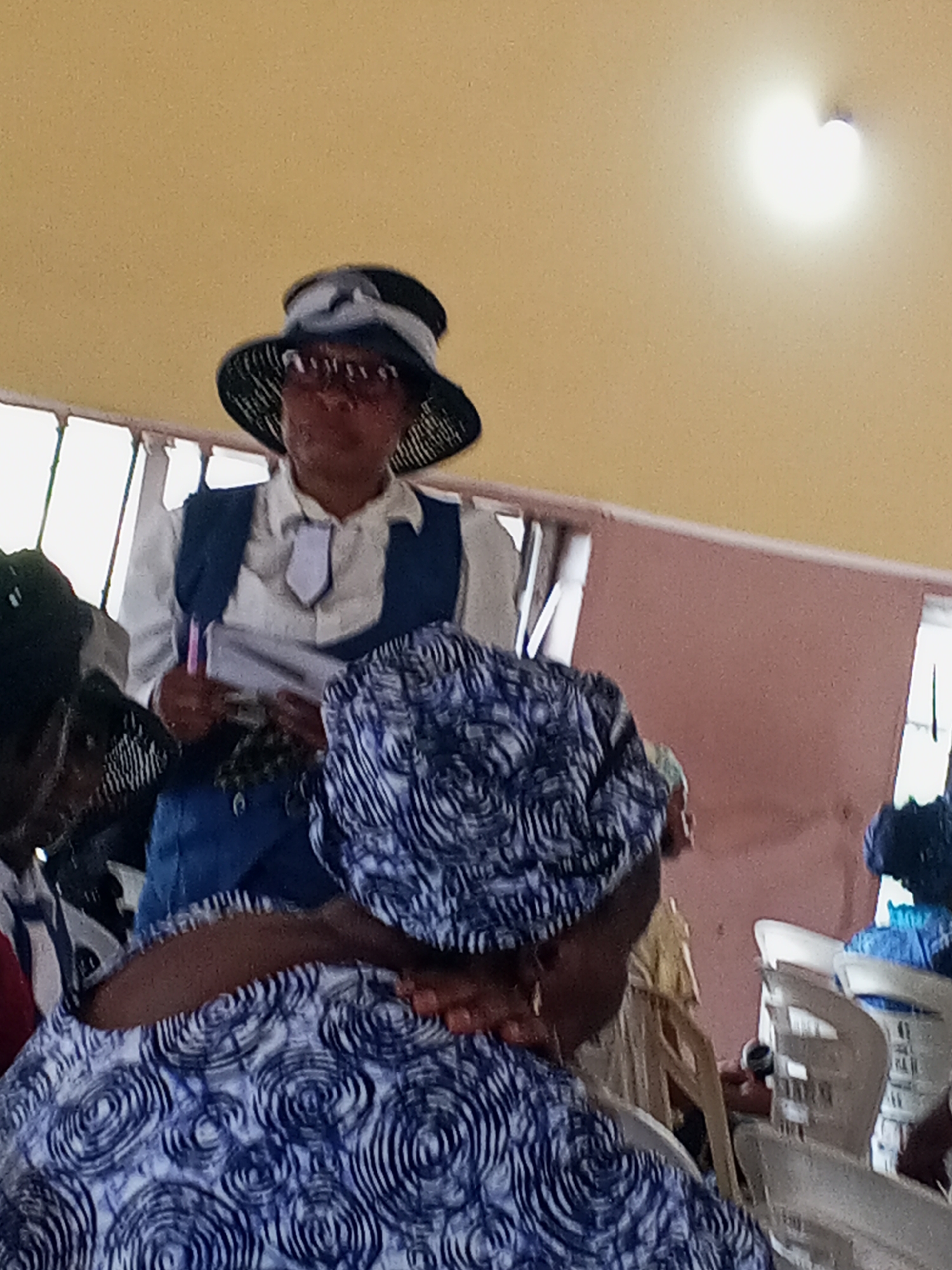 The image appears to be taken indoors, possibly in a church or meeting hall in Zango-ndaji, Nigeria. The ceiling is painted yellow with a bright light fixture. In the foreground and center of the image, several individuals are seated, with one person wearing a blue and white patterned headwrap. In the background, a woman stands dressed in a white shirt, a blue vest, and a black hat adorned with a white ribbon. She is holding a pen and papers. Other individuals are seated behind the person with the headwrap, and rows of white plastic chairs are visible. A window with white frames is visible in the background. The overall impression is one of a meeting or church service in progress.