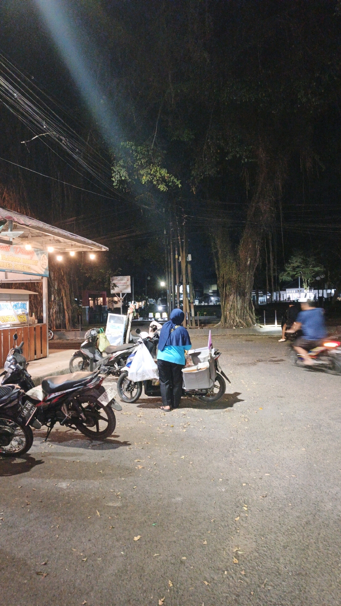 In the city of Desa Karangbenda, Indonesia, at night, a street scene unfolds with a focus on a food stall and its surroundings. A food stall with a white awning is visible on the left, displaying illuminated signs in Indonesian. Several motorcycles are parked along the roadside, some in front of the stall and others further down the street. A woman wearing a blue headscarf and a blue and teal outfit stands next to a motorcycle with a cart attached. She appears to be attending to the cart, which has a white bag on it. Another person is seated on a motorcycle further down the street, partially obscured by motion blur. In the background, a large tree with hanging vines dominates the scene, illuminated by artificial light. Streetlights and building lights are visible in the distance, suggesting a populated area. The ground is paved asphalt, with scattered leaves and debris. A bright beam of light cuts across the upper left portion of the image, likely from a street lamp or a vehicle. The overall atmosphere is that of a typical night market or roadside dining area in a local Indonesian community.