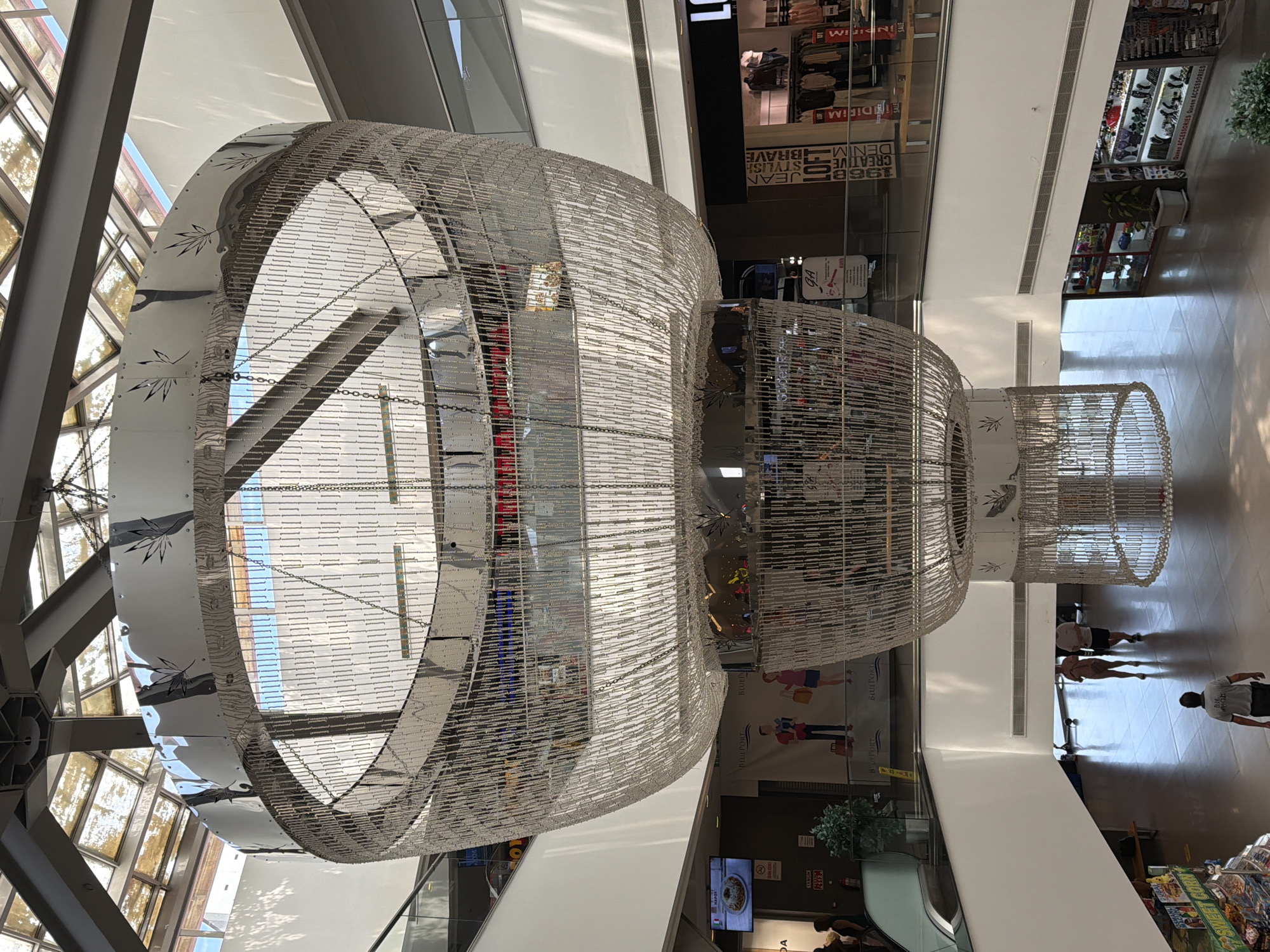 The image captures the interior of a multi-level shopping mall, likely the Marmaris Grand Bazaar in Marmaris, Turkey, focusing on a massive, multi-tiered chandelier that dominates the central atrium. The chandelier is an elaborate construction of metal and what appears to be beaded or chain-like material, suspended from the ceiling by chains. It has a distinctive, organic shape, widening as it descends and tapering towards the bottom. The upper section of the chandelier features a decorative band with a leaf-like pattern.
Sunlight filters through a glass roof above, illuminating the space and casting shadows. The mall's architecture is visible, with balconies and escalators on different levels. On one of the lower levels, several shops are visible, some with displays of clothing and accessories. A large screen displaying food is mounted on a wall.
There are a few people present in the scene. In the foreground, a man wearing a grey t-shirt and dark shorts is walking away from the camera, towards the bottom of the image. On a lower level, two women are walking together, and further back, a man is standing. The overall environment suggests a bustling commercial space.