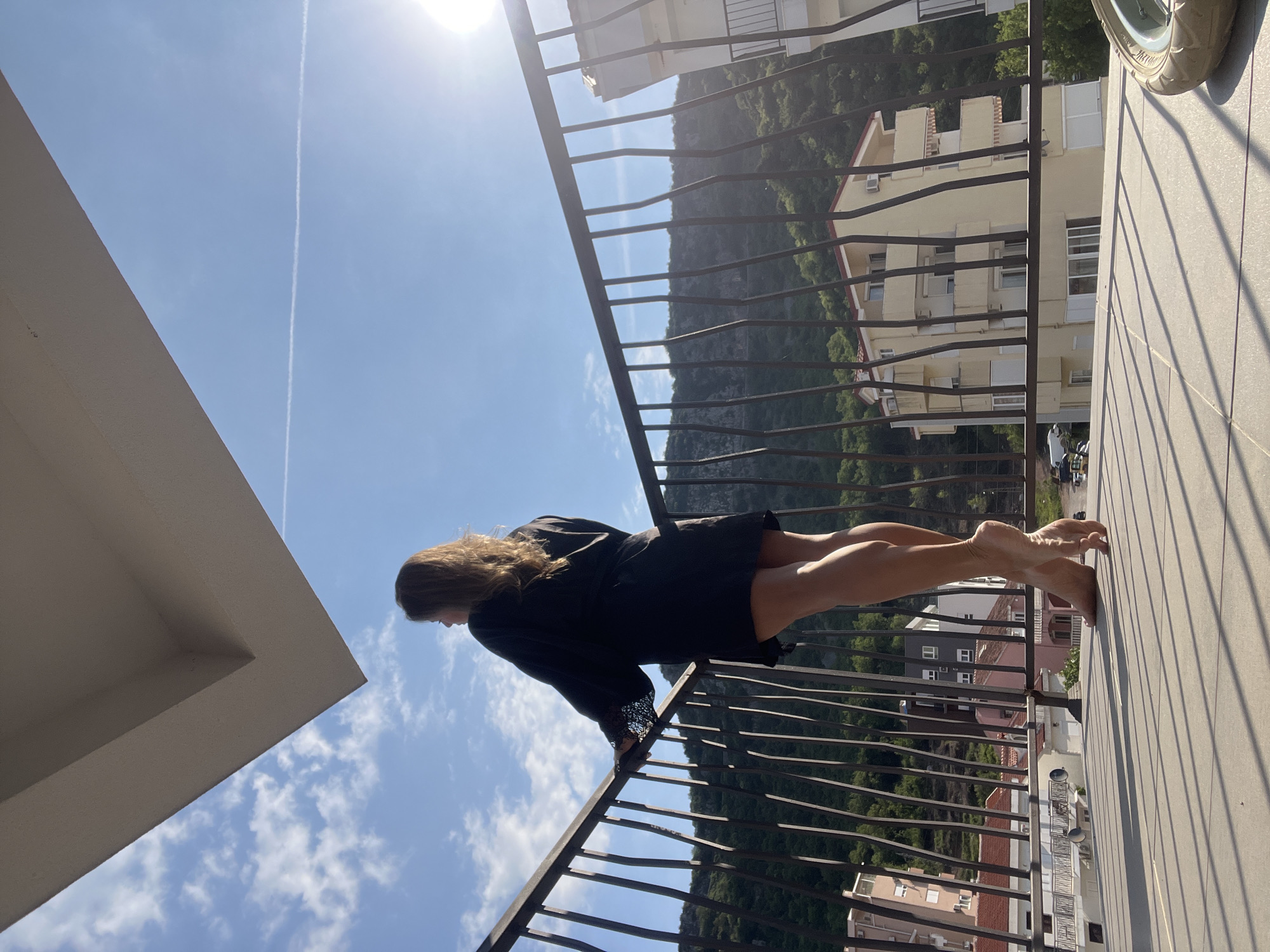 An outdoor, eye-level shot captures a woman on a sunny balcony overlooking a mountainous landscape and town. The provided image is rotated 90 degrees clockwise from its natural upright orientation.
The main subject is a woman with long, wavy brown hair, seen from behind and the side. She wears a black dress or robe with intricate lace detailing on the sleeves and is barefoot. She stands and leans against a dark metal balcony railing, with her body slightly arched and her feet pointed, looking out towards the panoramic view.
The scene features a bright blue sky with a few wispy white clouds and a distinct, long white contrail streaking across it. The sun is high and bright, casting strong shadows from the balcony railing onto the tiled floor. In the background, lush green mountains rise, behind which a cluster of multi-story residential buildings with light-colored facades, multiple windows, and balconies are visible. The architecture suggests a European, possibly Mediterranean, setting, consistent with the given location context of Budva, Montenegro. Part of a grey wheel, possibly from a scooter or motorcycle, can be seen on the far right of the tiled balcony floor. The overall impression is one of relaxation and admiration of the scenic vista on a clear, bright day. There is no clearly visible text in the image.