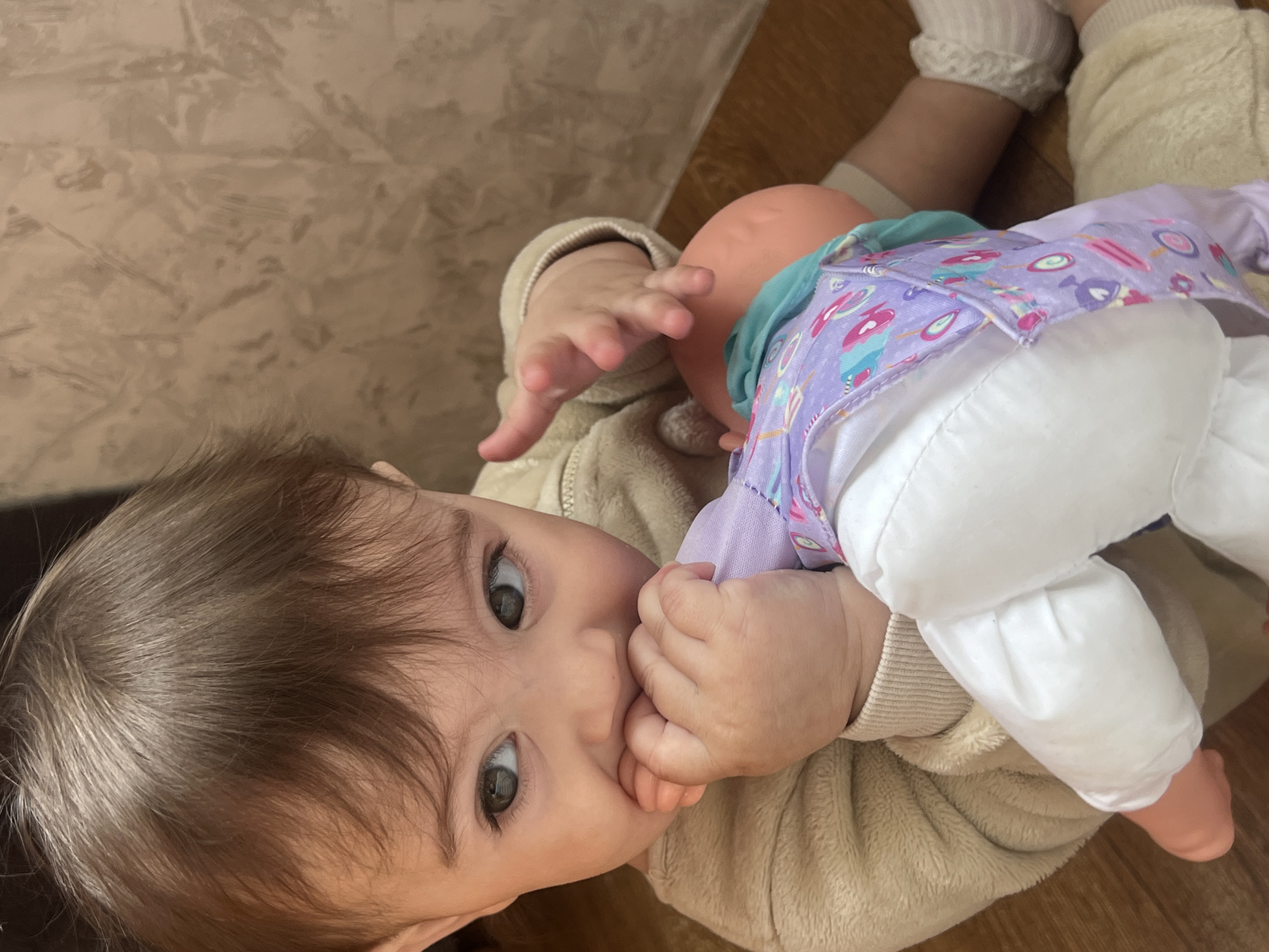 An infant or toddler with light brown hair and dark eyes is depicted in a medium shot, seated on a wooden floor in the city of Elena, Bulgaria. The child is wearing a light beige, plush long-sleeved garment and is holding a doll. The child's left hand is grasping the doll's hand, which the child is biting or chewing. The child's right hand rests on the doll's torso. The doll has a light pink plastic or rubber body and is dressed in a purple top patterned with colorful shapes (hearts, popsicles, circles) and white pants. The child is looking intently towards the left side of the frame. The background consists of a light brown wall with an abstract, textured pattern.
