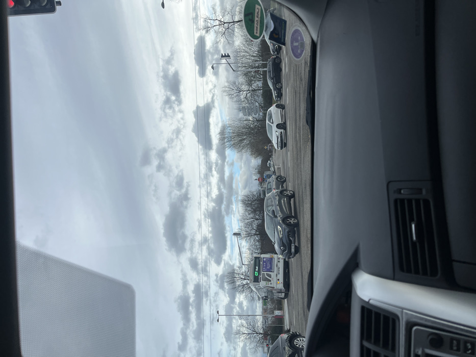 The image presents a forensic view from inside a vehicle at a multi-lane road intersection in Illkirch-Graffenstaden, France, at the crossing of Route de Lyon (D468) and Rue de l'Industrie / Avenue de Strasbourg. The weather is overcast with a mix of grey and blue sky.
The lower foreground is occupied by the dark grey dashboard of the observer's vehicle, featuring air vents and control panels. The upper part of the windshield shows a partial view of a red-illuminated traffic light on the right and several circular stickers adhered to the glass.
At the intersection, a dark grey Renault Captur SUV, bearing French license plate DL-463-MY, is centrally positioned. To its right, a white Volkswagen Scirocco is visible, followed by a black Mercedes-Benz C-Class sedan and another dark sedan further right. A black SUV is partially visible on the far left. A white public transport bus, marked with "C7 Sud" and "T'RIGA" branding, is situated in the left lane behind the Renault Captur. A green canopy, indicative of a gas station, is visible behind the bus. Bare trees, traffic lights, and streetlights line the intersection and surrounding areas. A distinctive tall, modern building, likely part of the Parc d'Innovation, is visible in the distant background. Vehicles are either stopped at the red light or preparing to move through the intersection.
